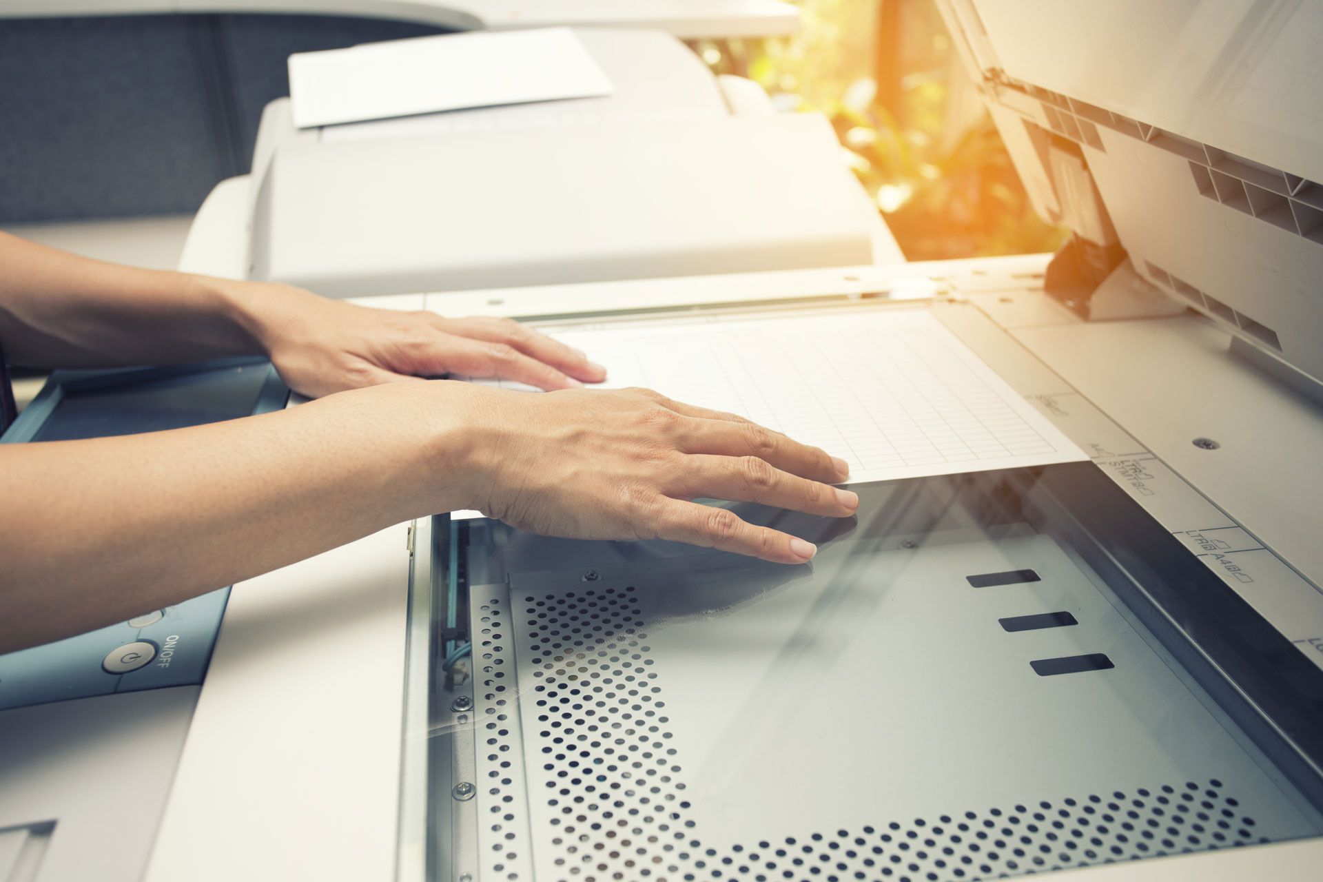 Hands pressing a sheet of paper onto the glass platen of an open office photocopier.