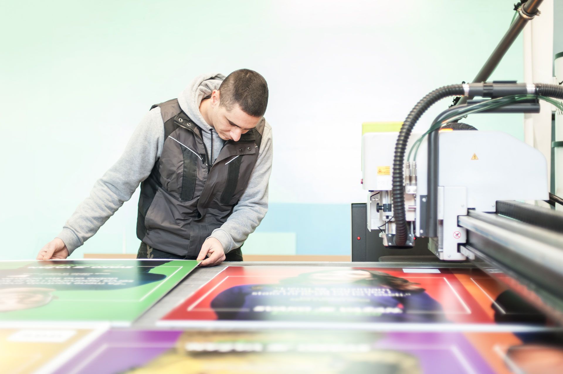 A person in a gray vest aligns a printed poster on a large industrial cutting machine in a workshop.