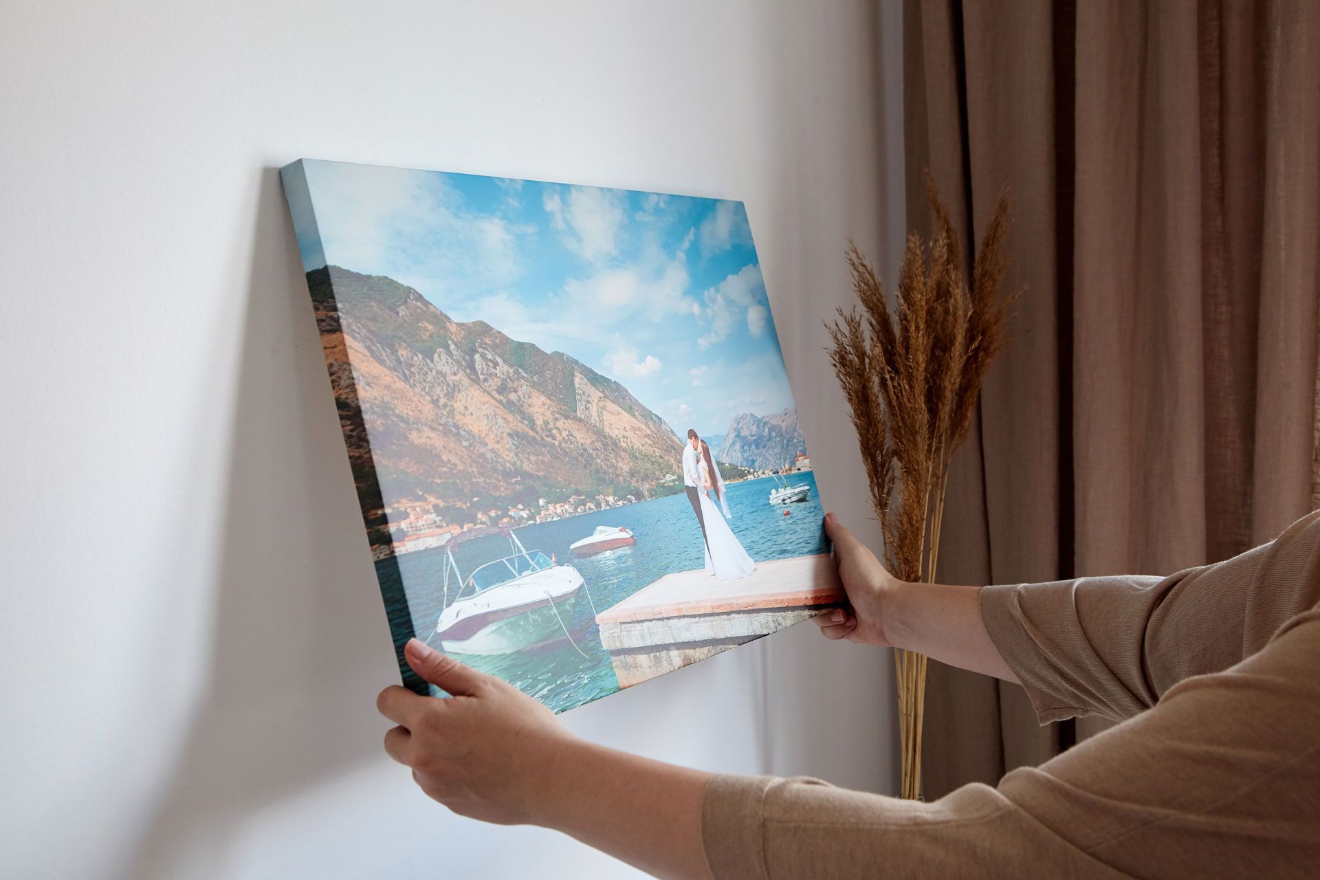 A person hanging a canvas print featuring a wedding photo by a lake and mountains on a white wall.
