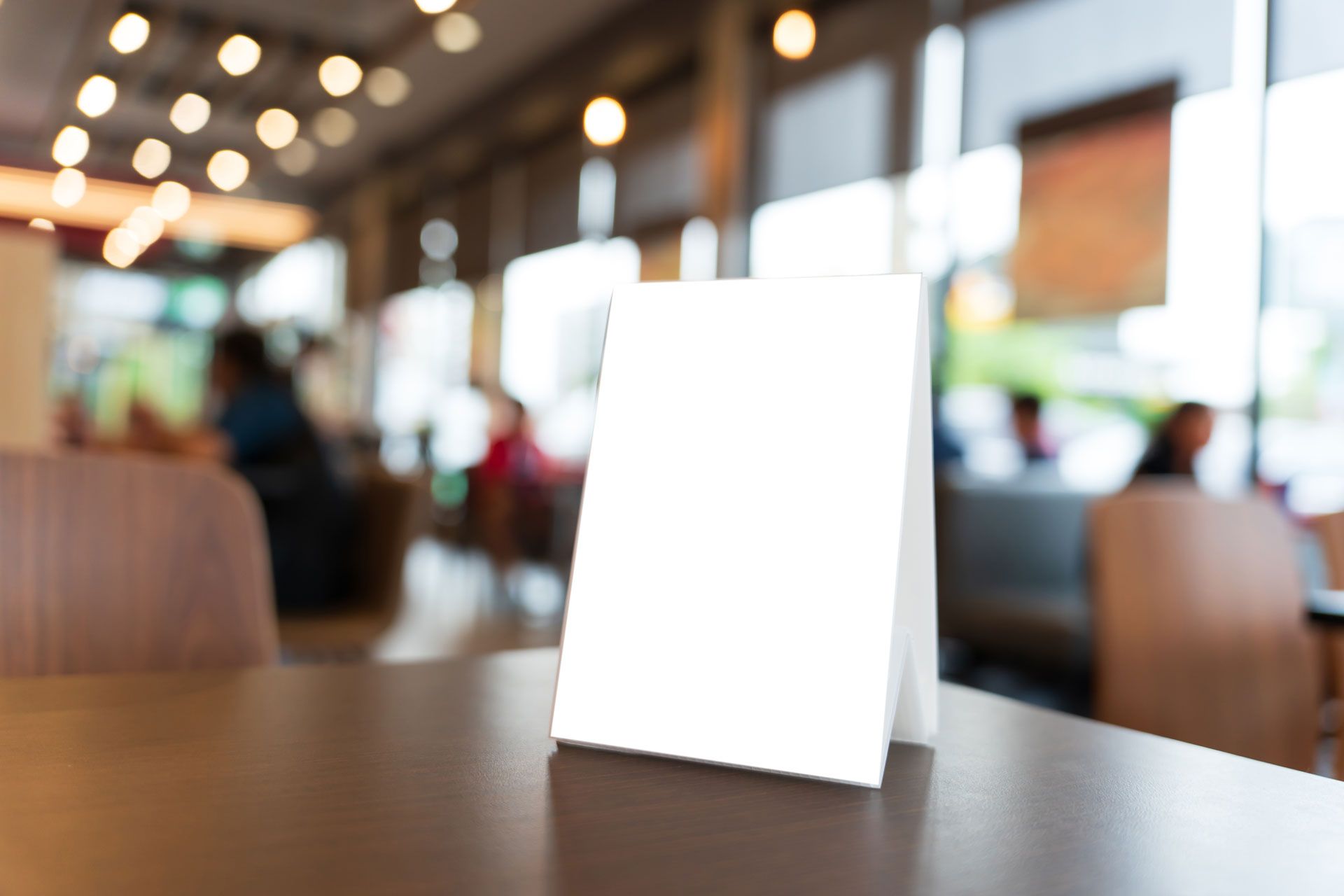 A blank white table tent card stands on a wooden table inside a softly blurred restaurant setting.
