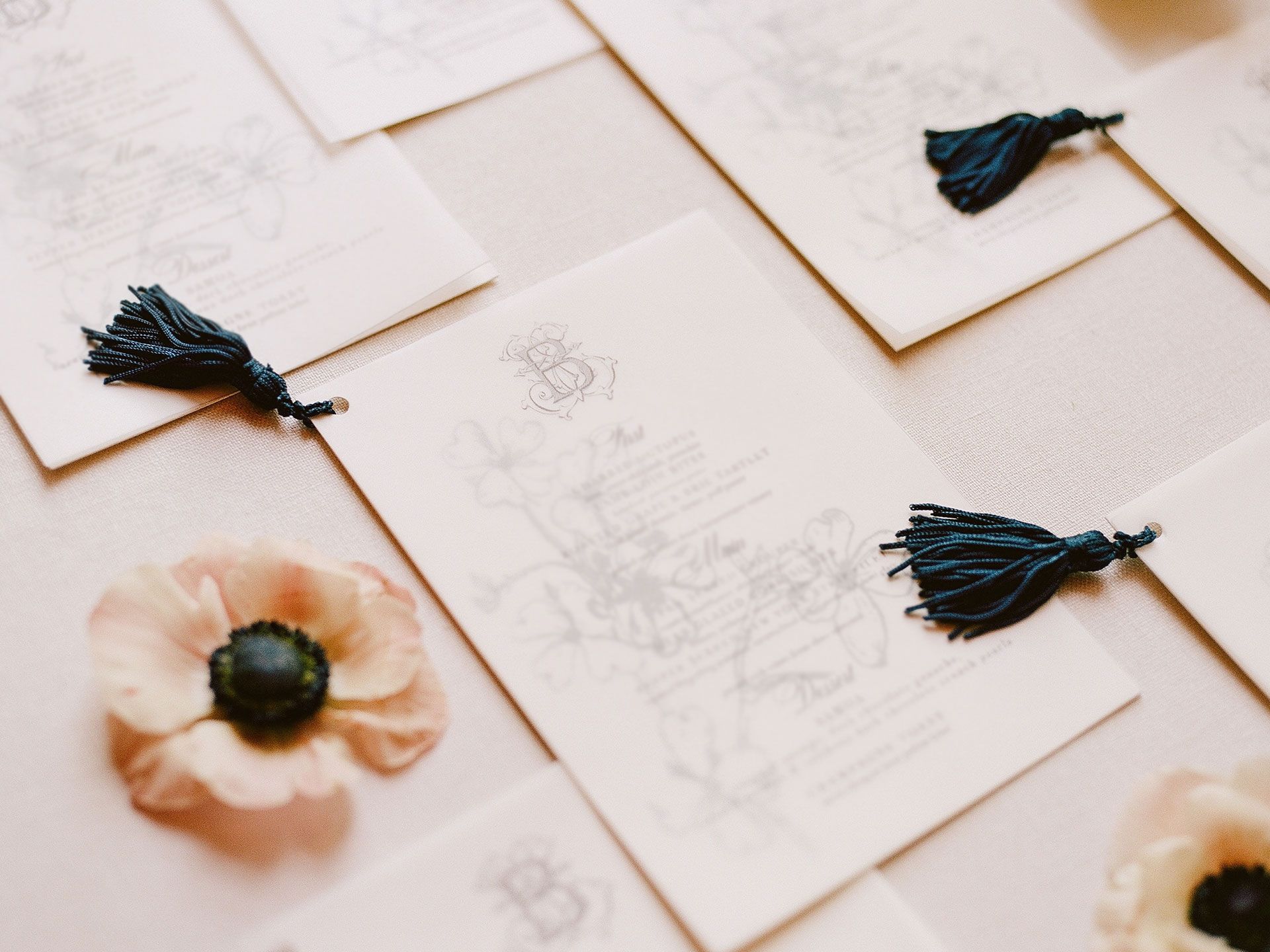Cream-colored paper cards with ornate floral sketches and dark blue tassels, accented by soft, pale flowers.
