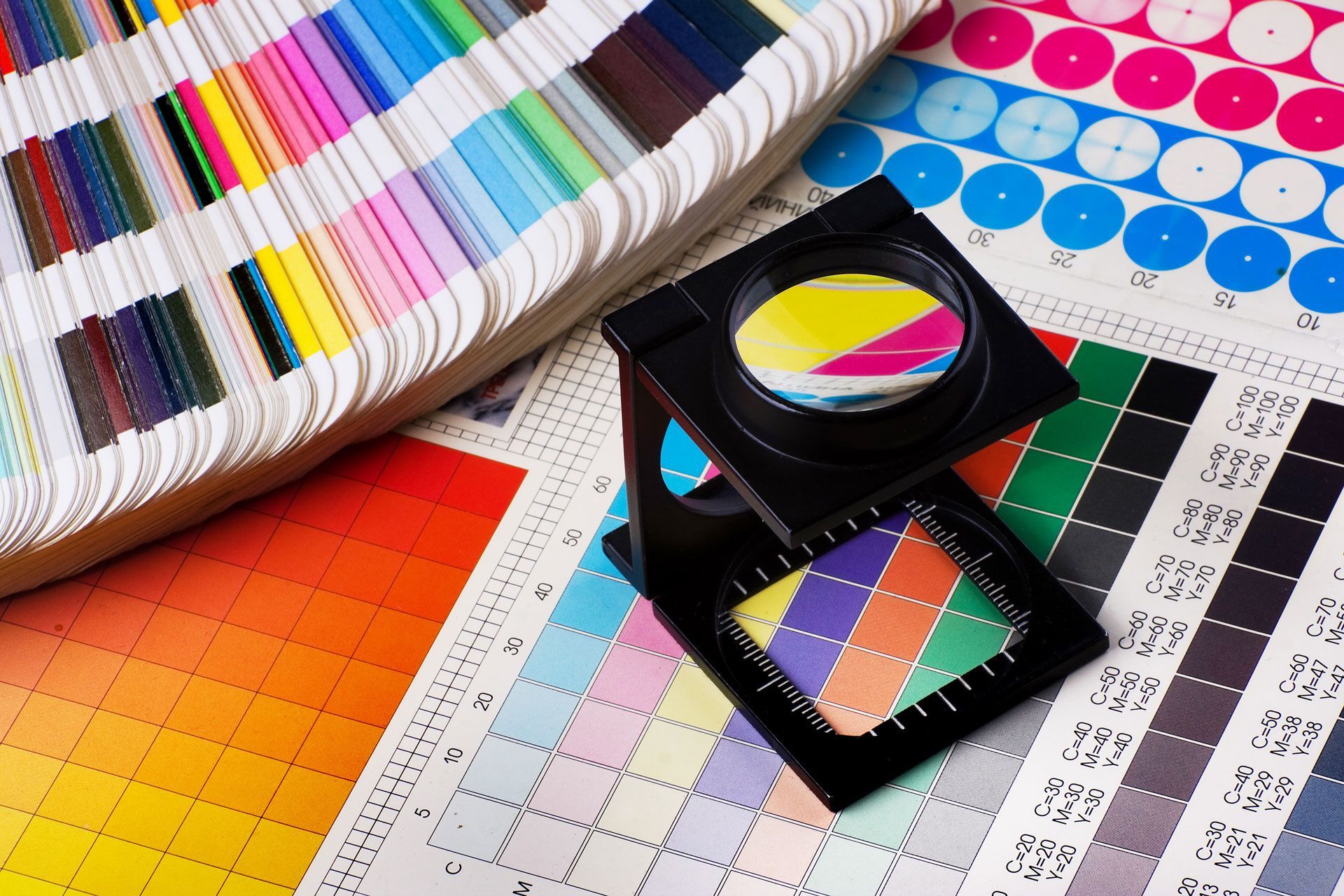 A printer's magnifying glass sits on color calibration charts and a Pantone swatch book for quality control.
