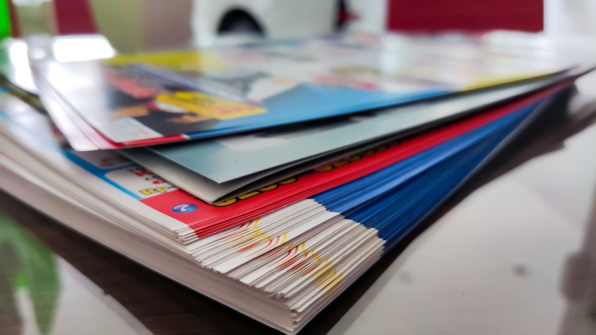 A stack of magazines and documents rests on a table, shown from a low angle with a shallow depth of field.