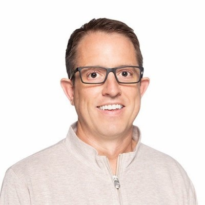 A smiling person wearing glasses and a light-colored, zippered top against a plain white background.