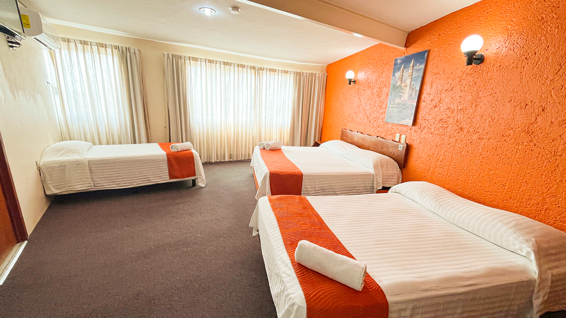 Hotel room with three beds featuring white linens and orange runners, against a textured orange wall.
