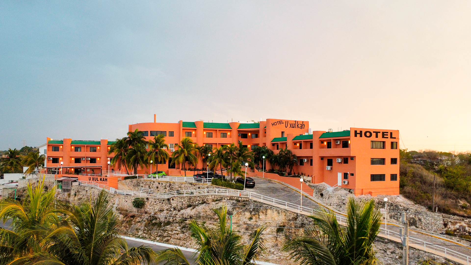 A multi-story, orange-colored hotel building set against a clear sky, situated above a rocky, palm-lined landscape.