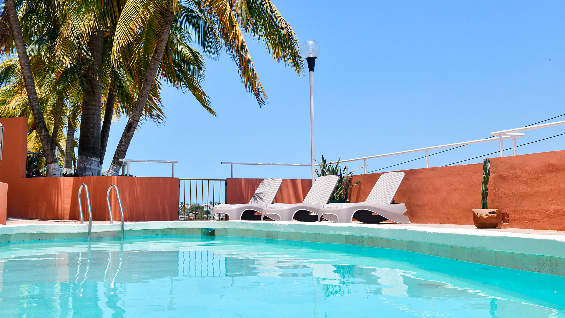 A bright blue swimming pool sits in front of three white lounge chairs against an orange wall under a clear blue sky.