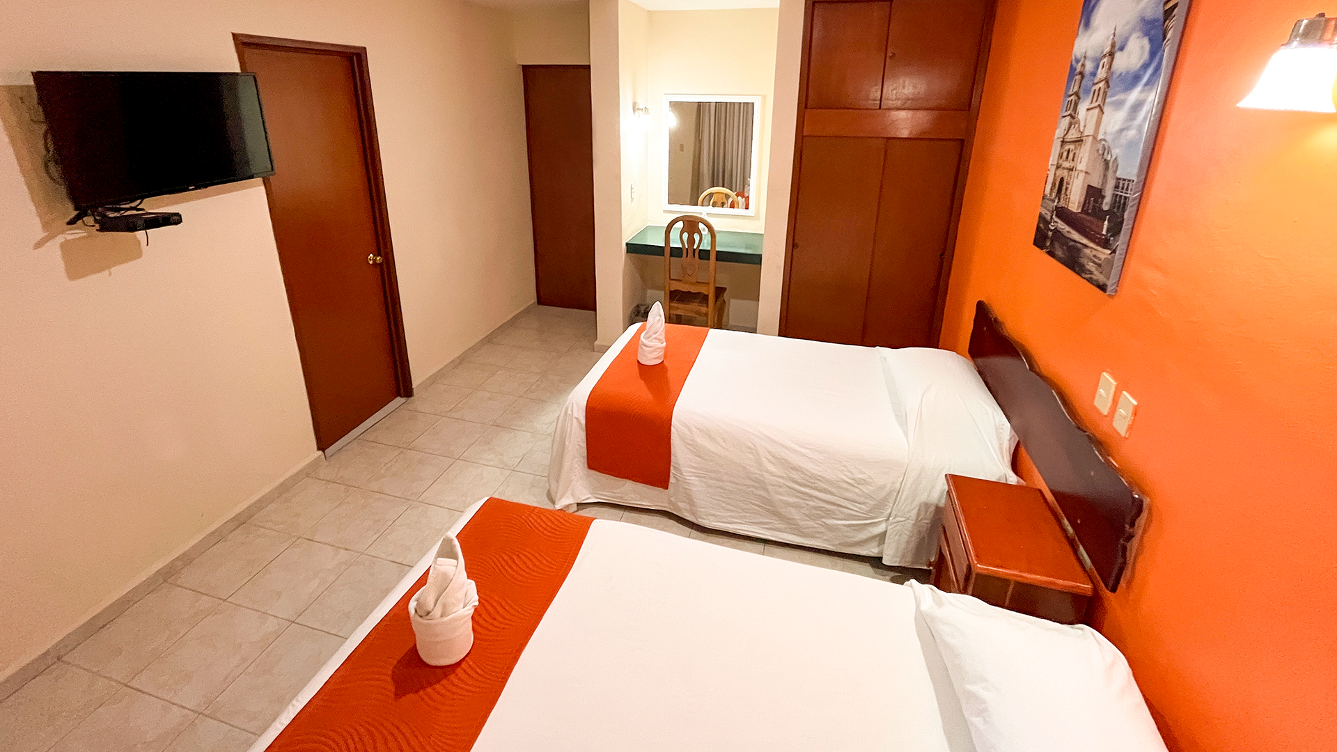A hotel room with two white beds featuring orange runners, a television on a wall, and an orange-painted wall with art.