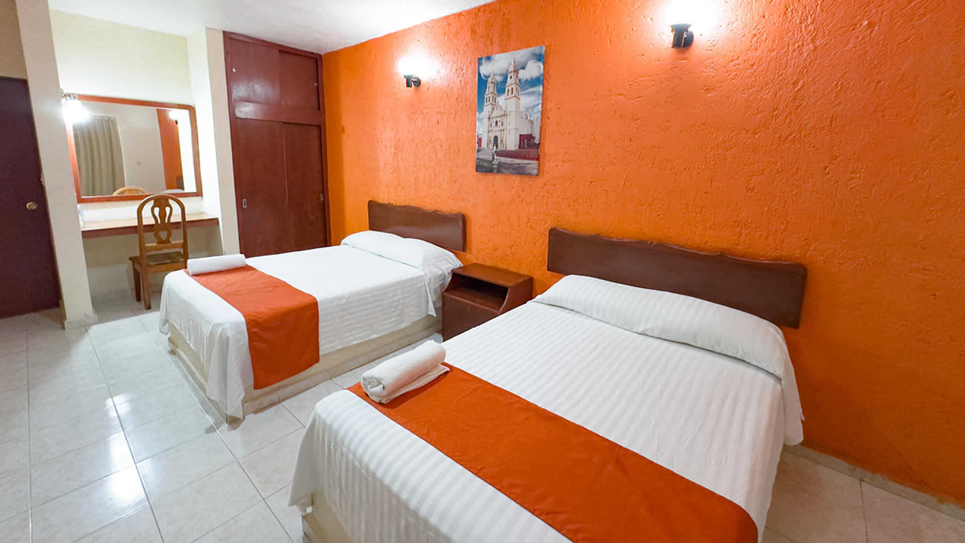 A hotel room with two beds featuring white linens and orange runners, set against bright orange textured walls.
