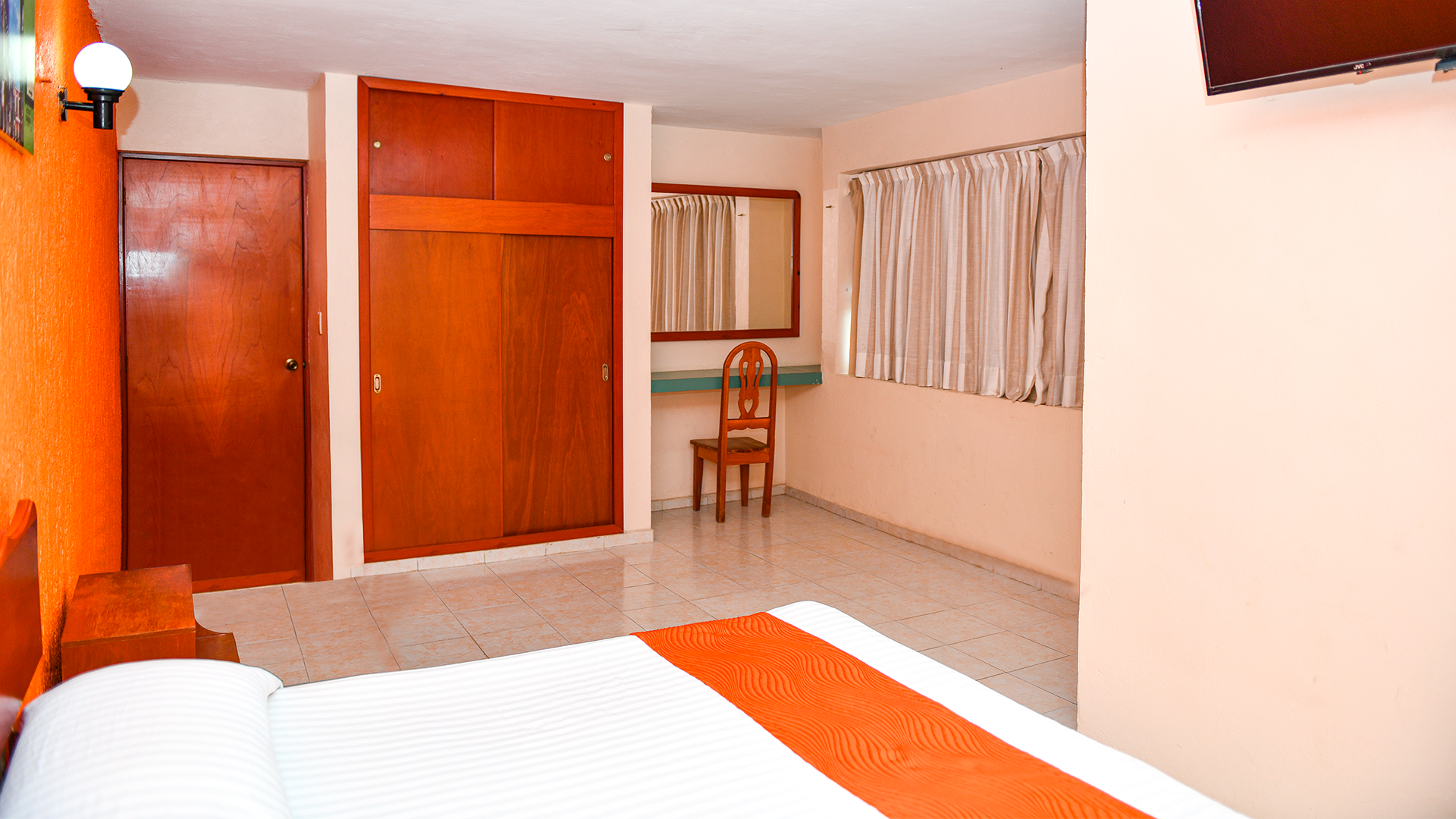 A hotel bedroom with a bed in the foreground, a wooden wardrobe, a desk with a chair, and a window with blinds.