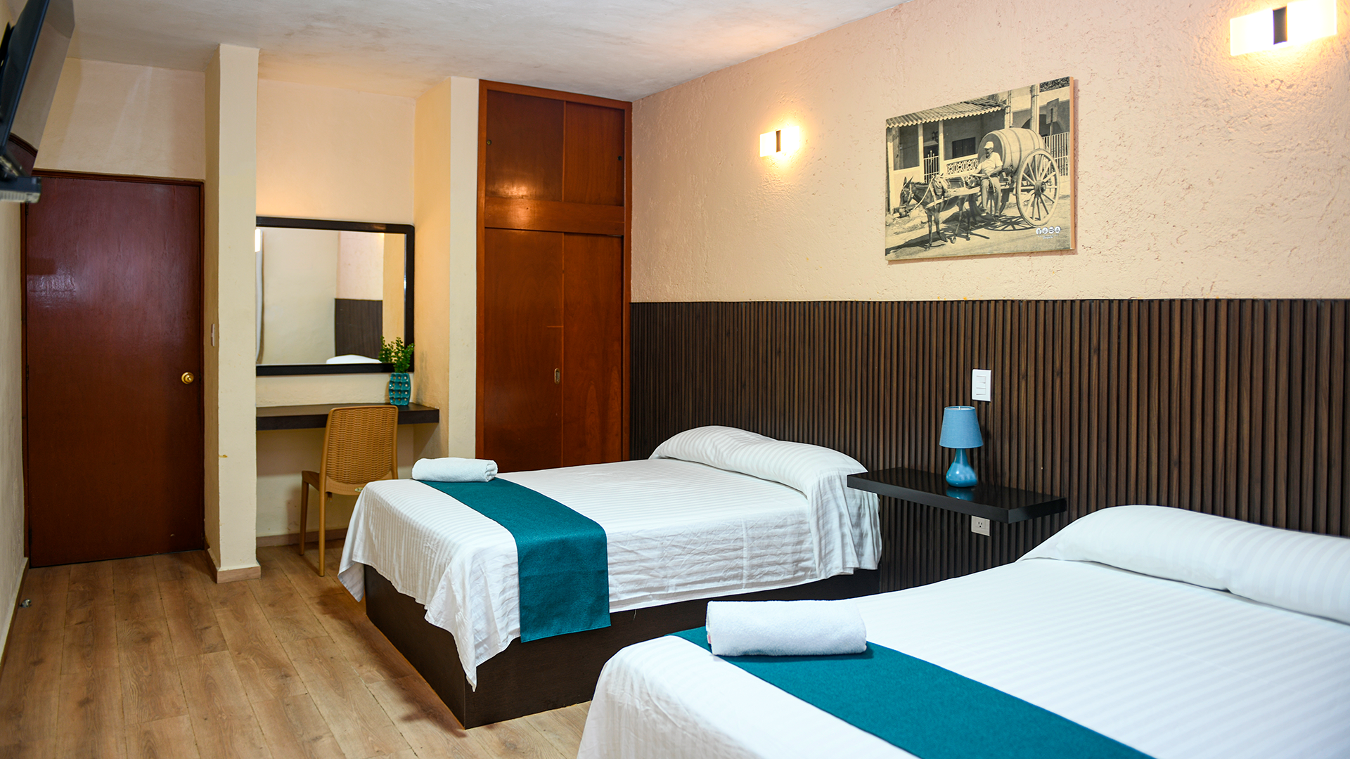 A hotel room with two beds featuring white linens and teal runners, a wooden desk, and a wall painting.