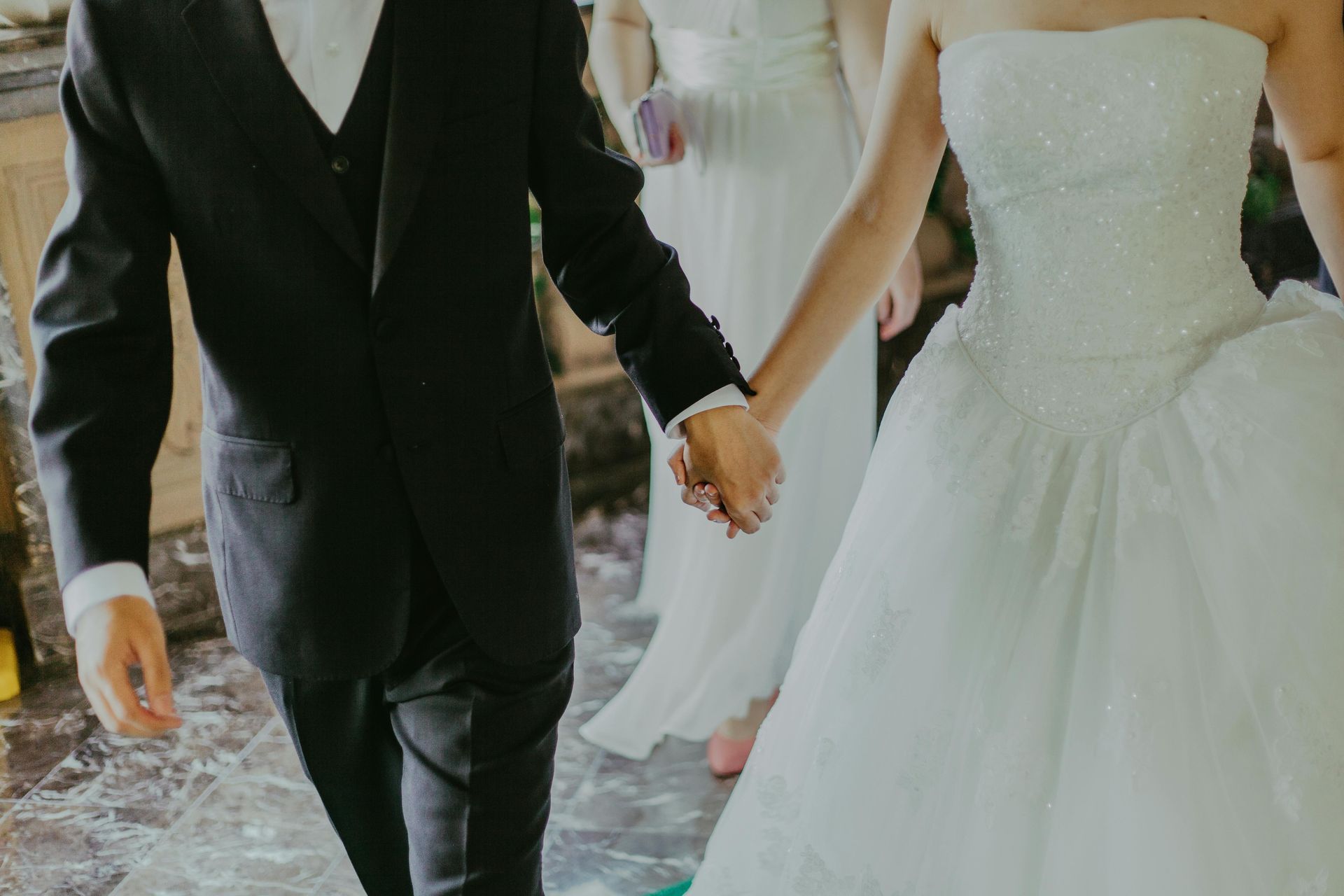 A person in a black suit holds the hand of another in a white wedding gown, with a second person in white in the background.