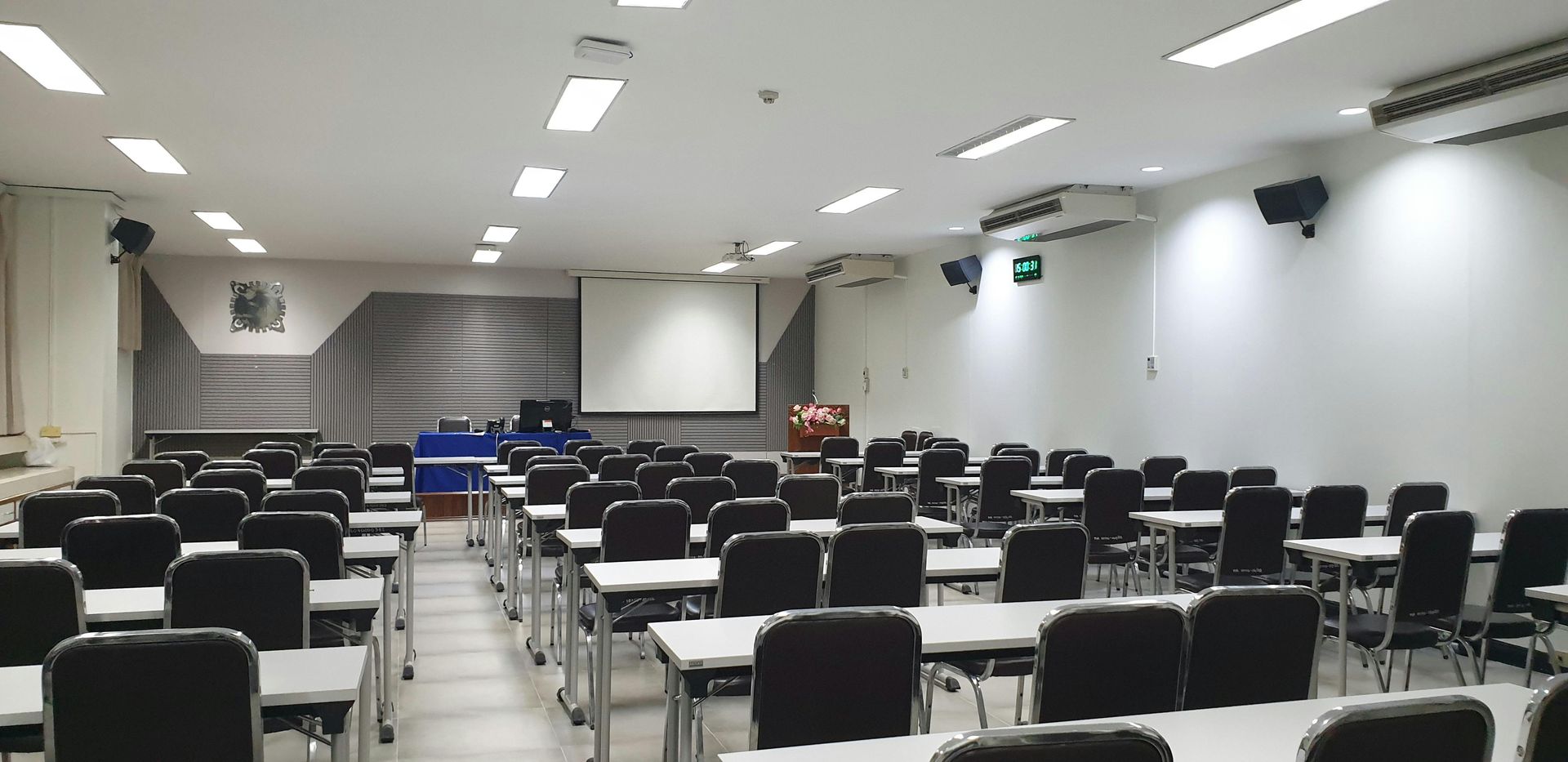 An empty classroom featuring rows of desks and black chairs facing a projector screen at the front.