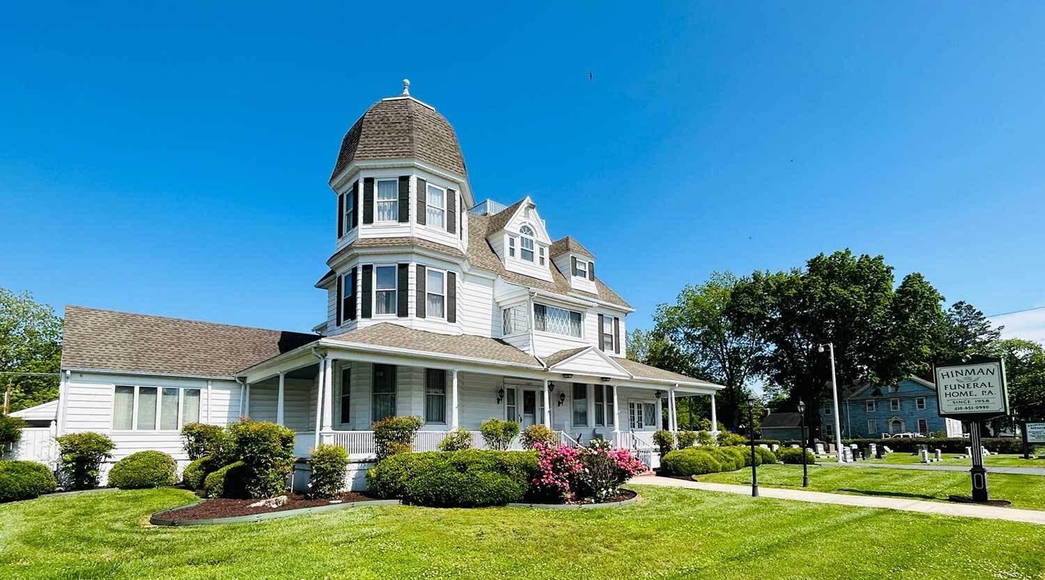White Victorian house with dome-shaped tower, porch, and manicured lawn under a blue sky.