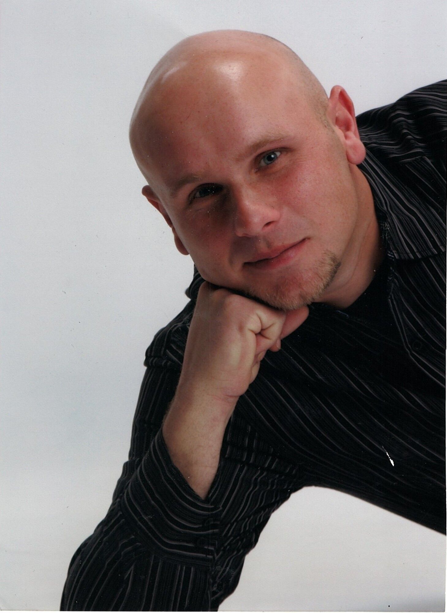 Bald man with hand on chin, smiling, wearing a striped shirt, leaning on a surface, against a white background.