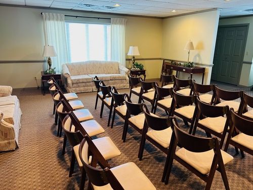 Rows of chairs face a sofa in a room, likely a chapel or waiting area with lamps and a window.