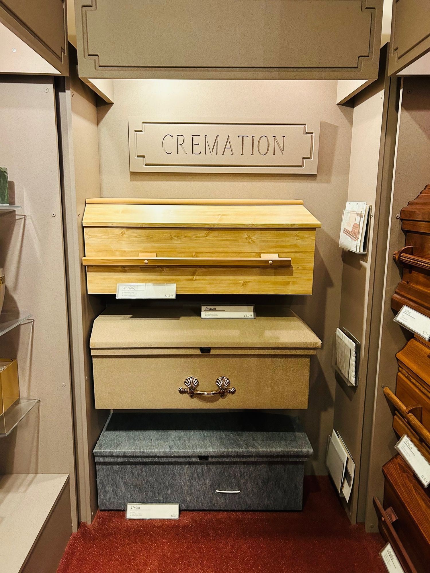 Cremation caskets displayed in a funeral home setting. Wooden, beige, and gray options are shown.