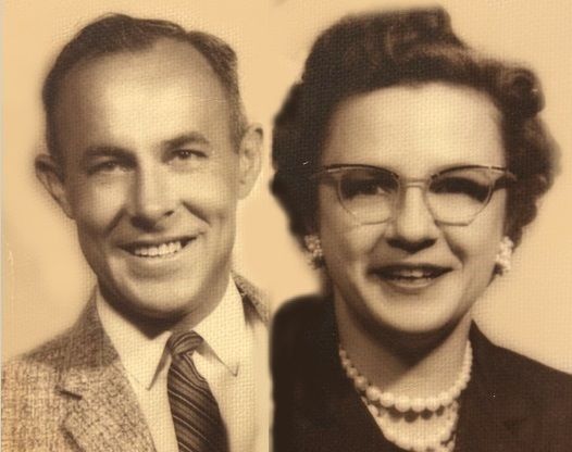 Couple smiling at the camera. Man in a suit and tie, woman with glasses and pearls.
