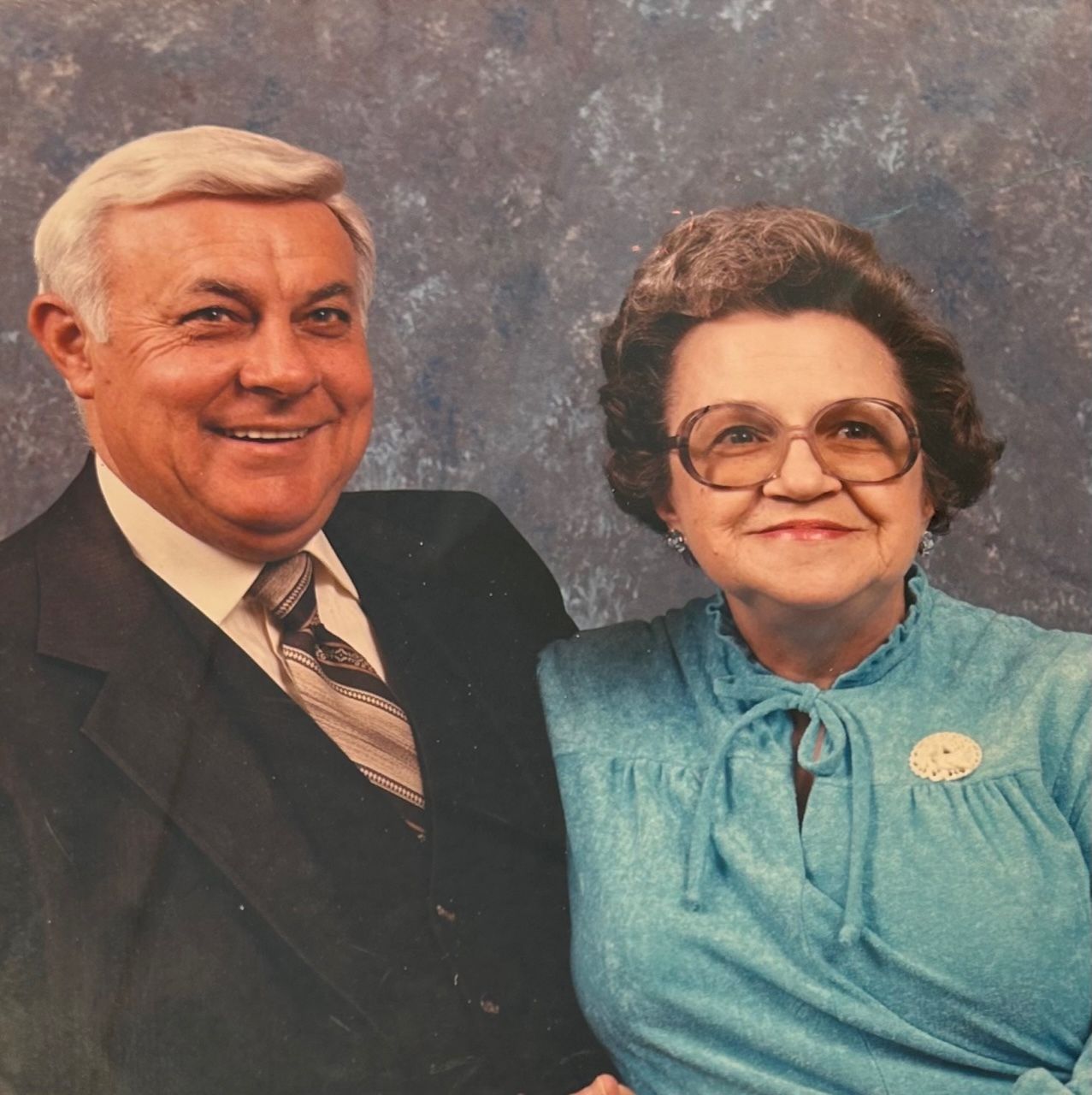 A smiling man in a suit and a woman in a blue blouse pose together in a studio setting.