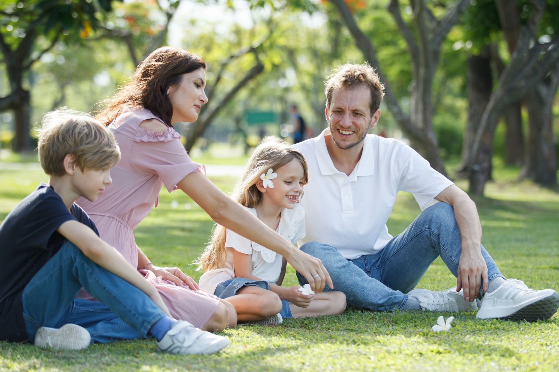 Family sitting on grass in a park, smiling and interacting, under a tree.