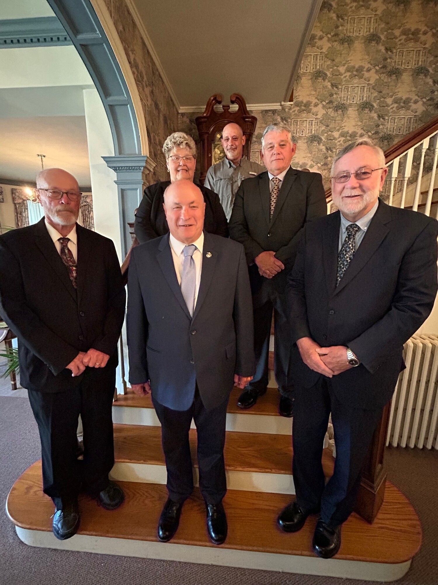 Group of six people posing on steps, indoors. Men wear suits. A woman is in the middle row. They face forward.