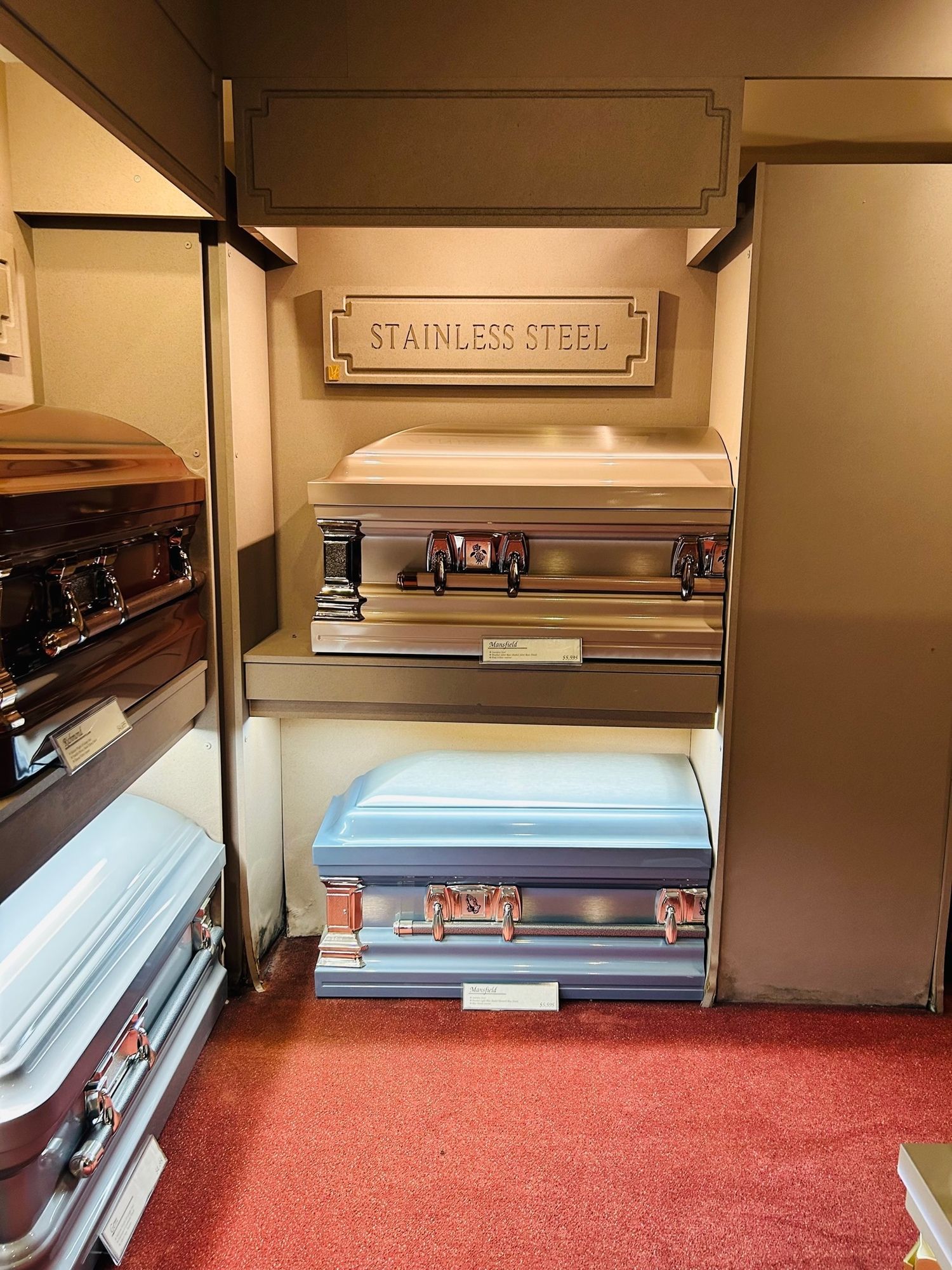 Inside a funeral home showroom, stainless steel caskets displayed on shelves with a red carpet.