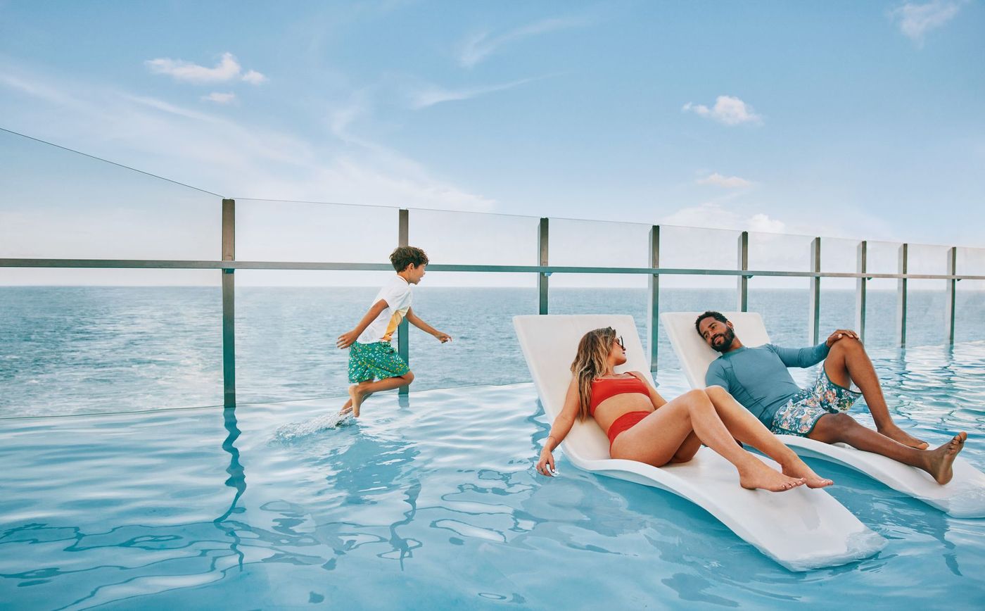 A man , woman and child are sitting on lounge chairs in an infinity pool overlooking the ocean.