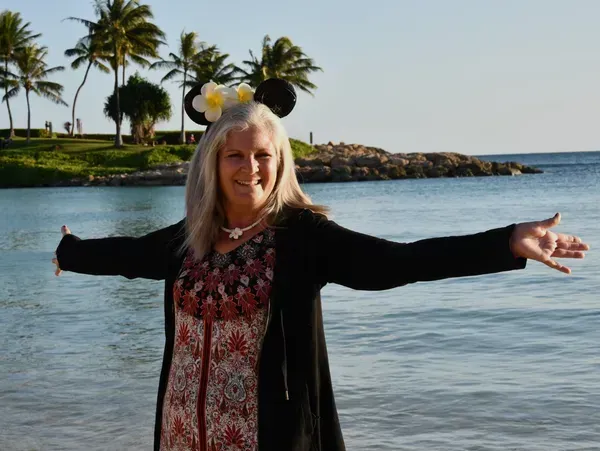 A woman wearing mickey mouse ears stands on the beach with her arms outstretched