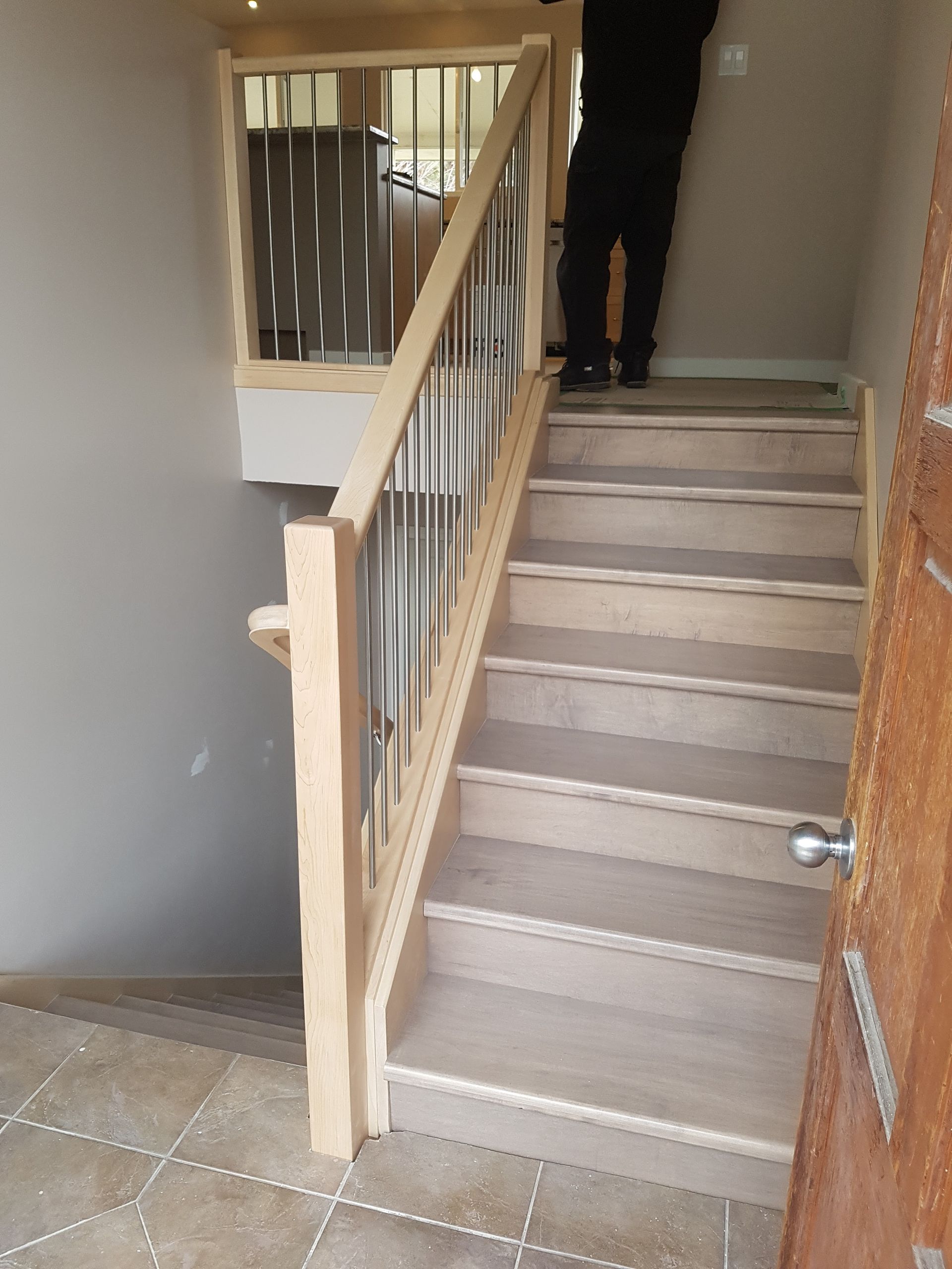 Staircase with wooden banister and stainless steel bars. A person stands at the top of the stairs near a door.
