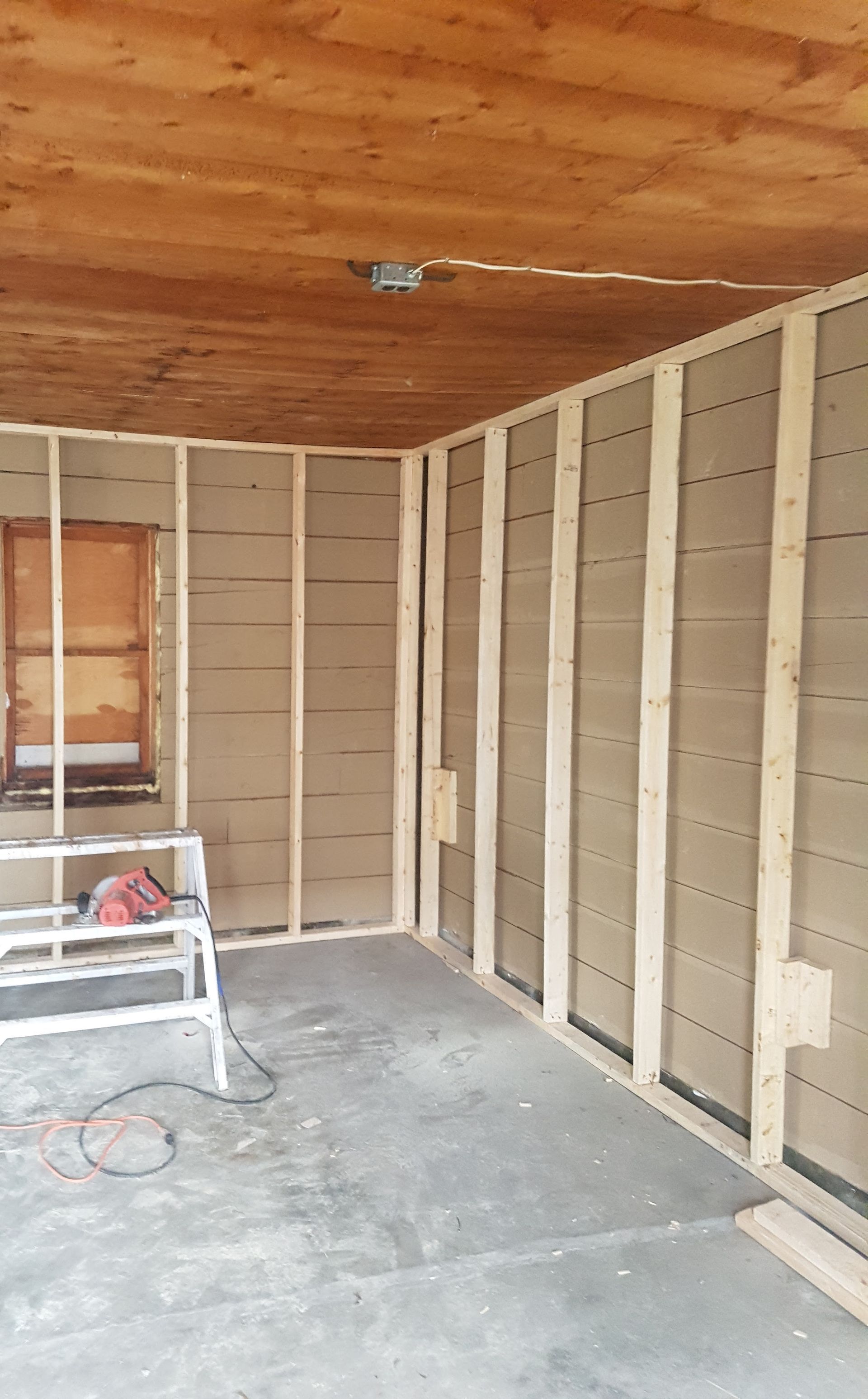 Interior of a room with wooden framing and insulation, unfinished concrete floor, and a partially visible window.