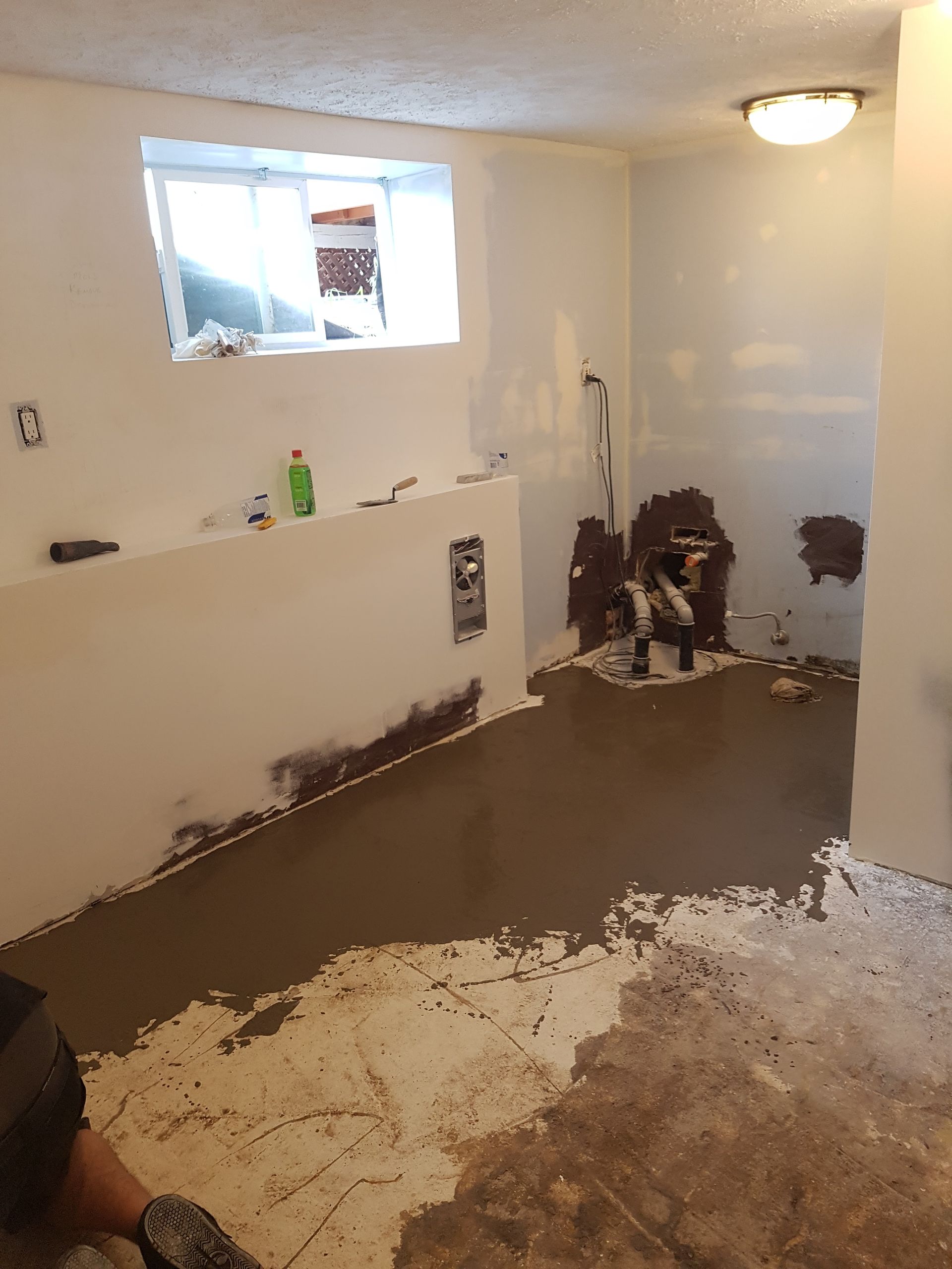 Basement renovation in progress: gray concrete floor, white walls, person working near pipes, window.