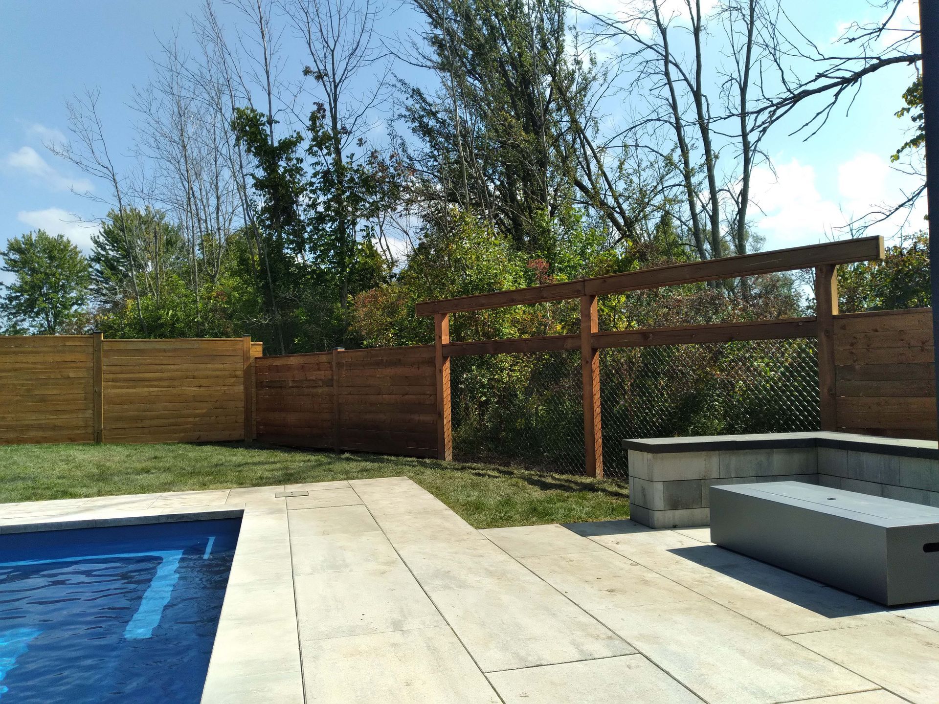 A backyard with a pool, patio, and wooden fence under a blue sky.