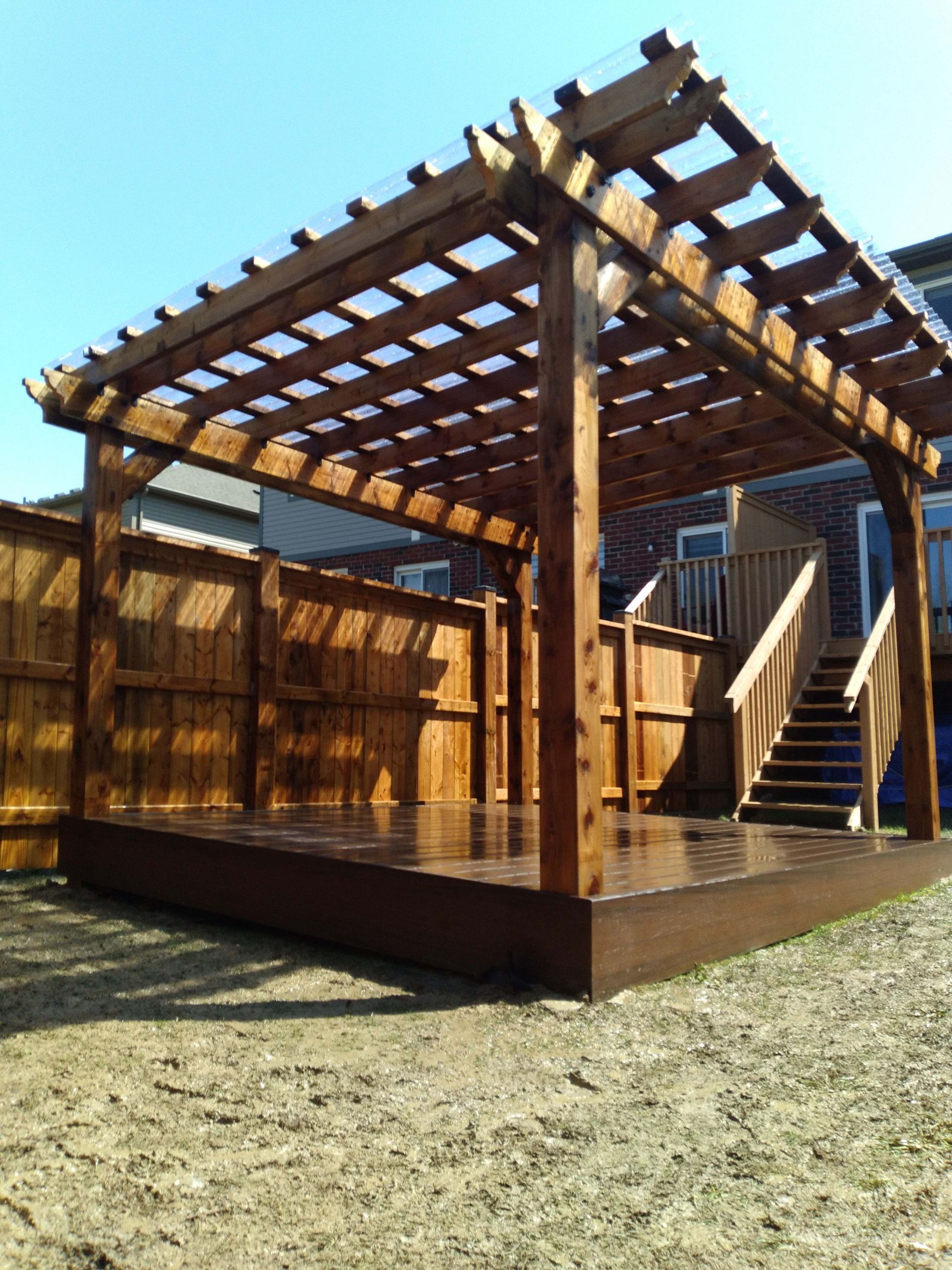 Wooden pergola over a deck with stairs, brown stain, clear sky, surrounded by a fence.