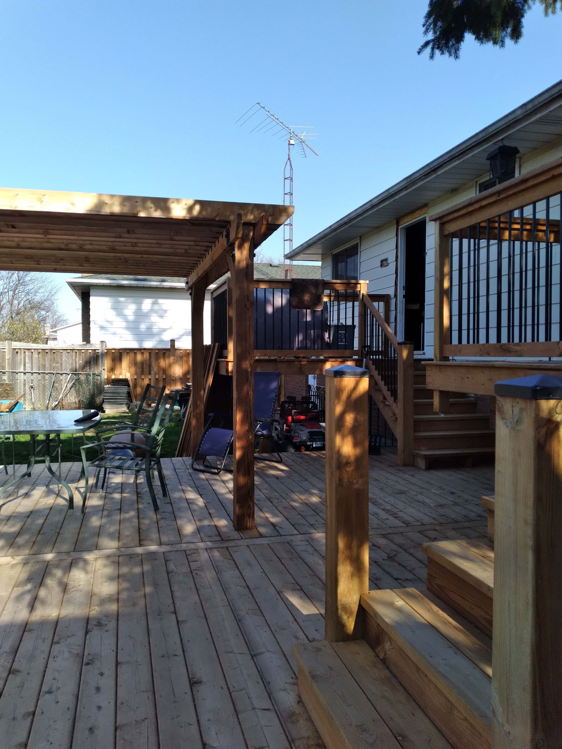 Wooden deck with pergola, leading to a house with a small yard and an antenna. Sunny day.