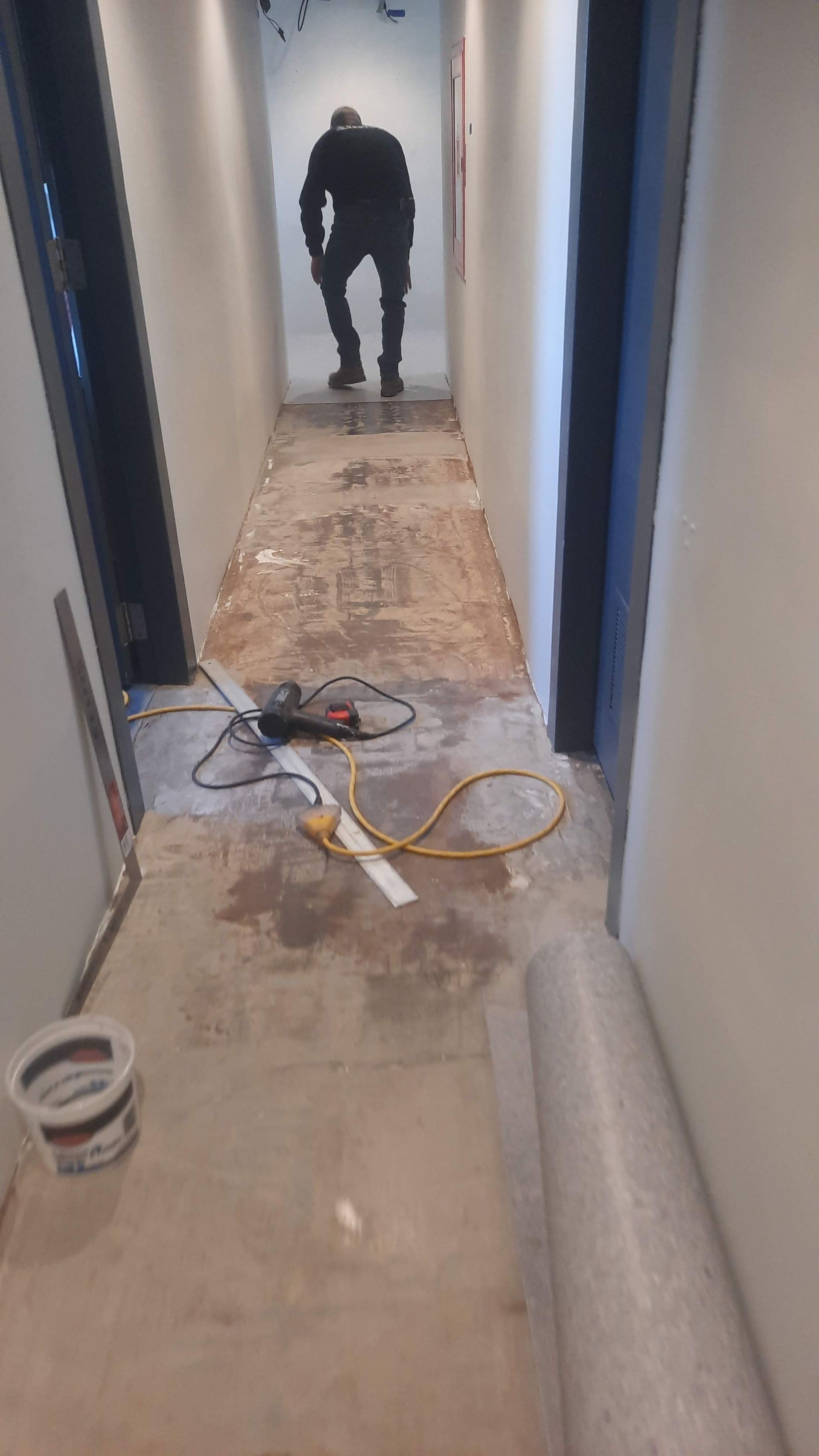 Narrow hallway under renovation; man walking towards end. Concrete floor with debris, tools, and rolled material.