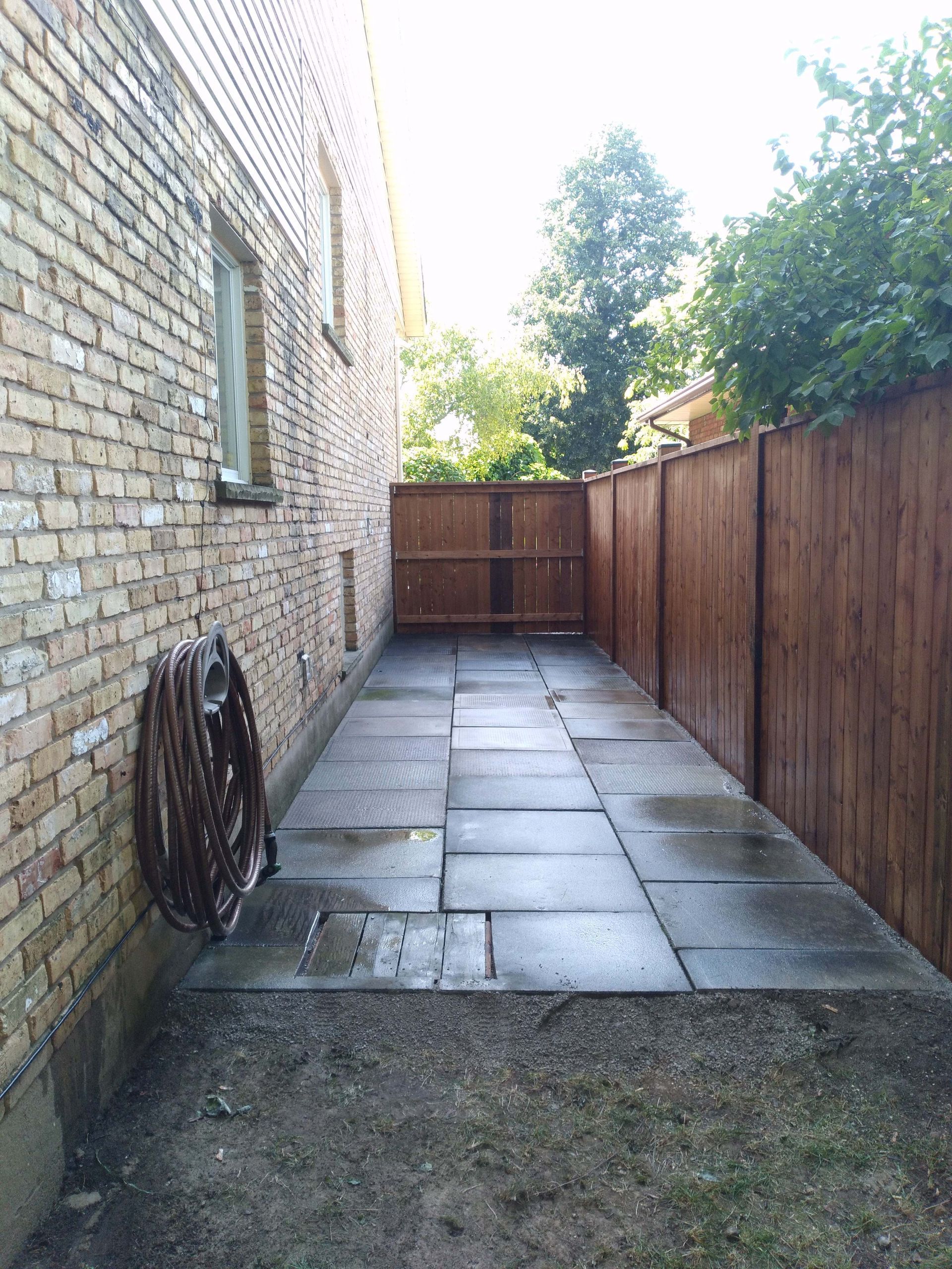 Brick building side with stone patio, brown fence, coiled hose, and green grass.