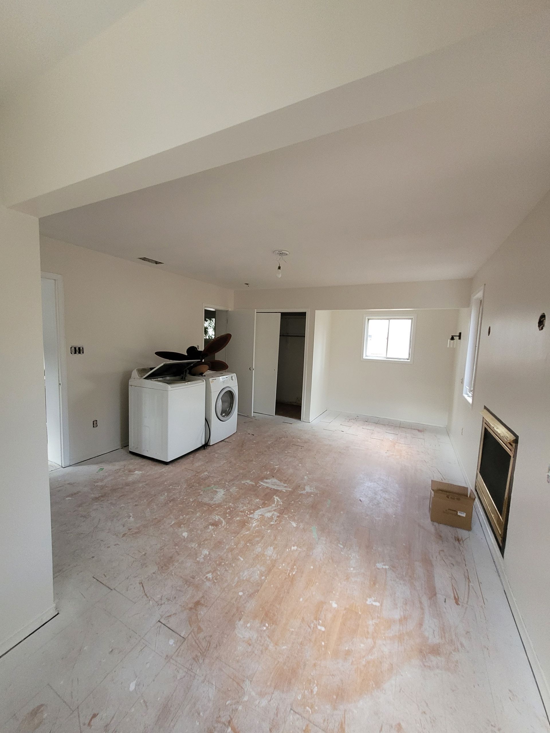 Empty room undergoing renovation, white walls, bare wood floor, washer/dryer visible, doorway, small window, and fireplace.