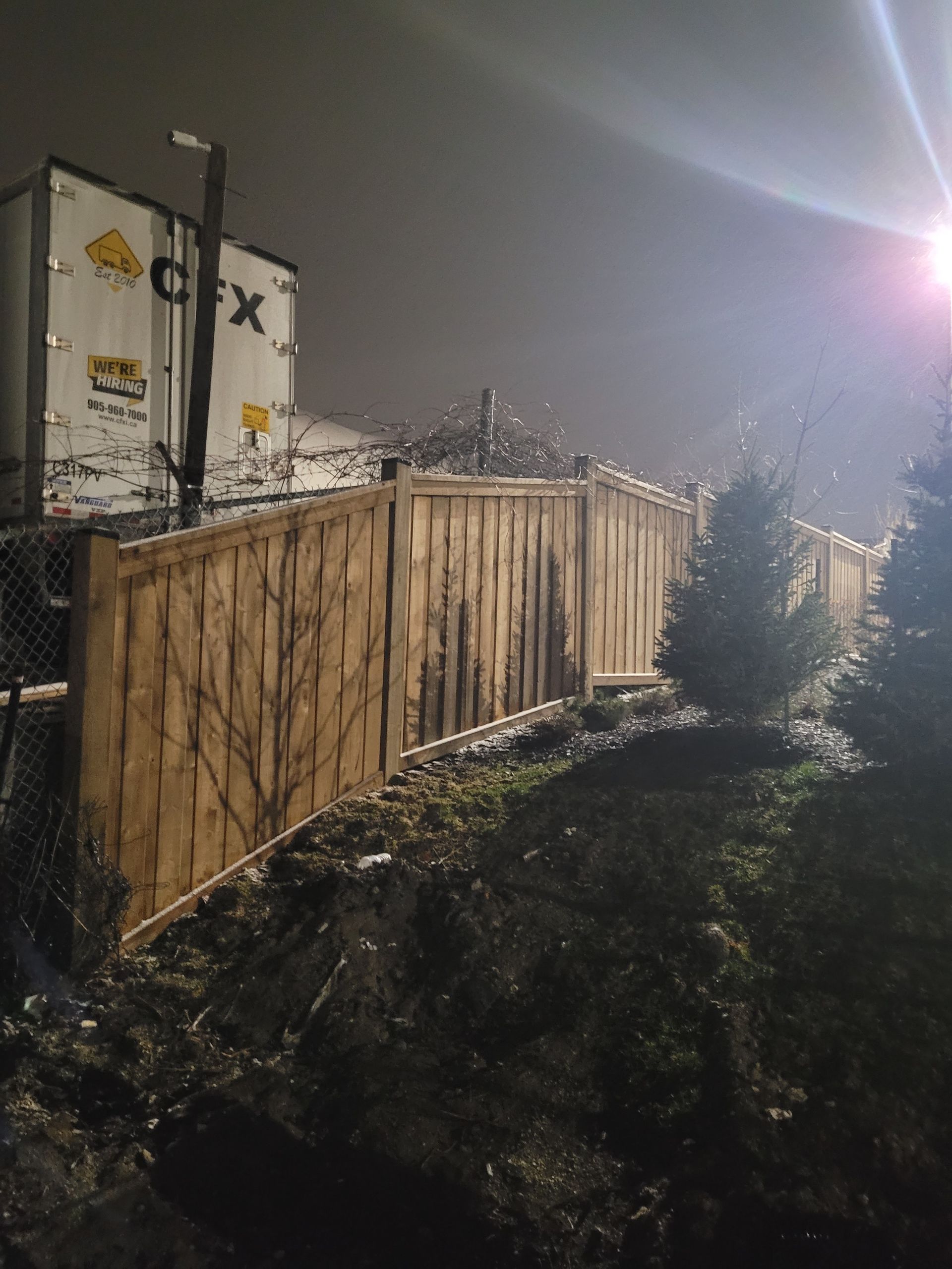 Wooden fence and cargo containers at night, illuminated by a bright light.