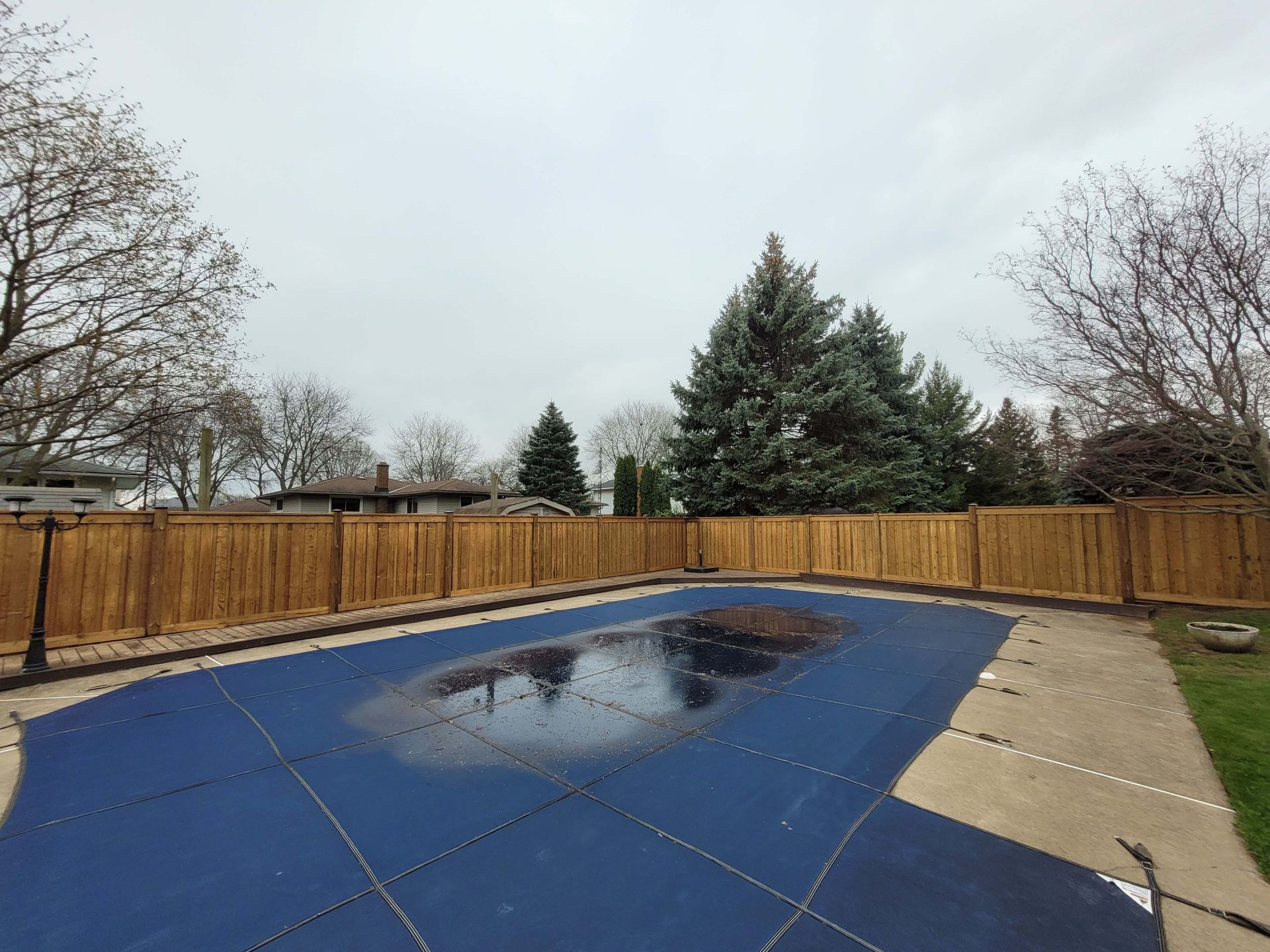 Pool covered in blue tarp, surrounded by a wooden fence and green grass under a cloudy sky.