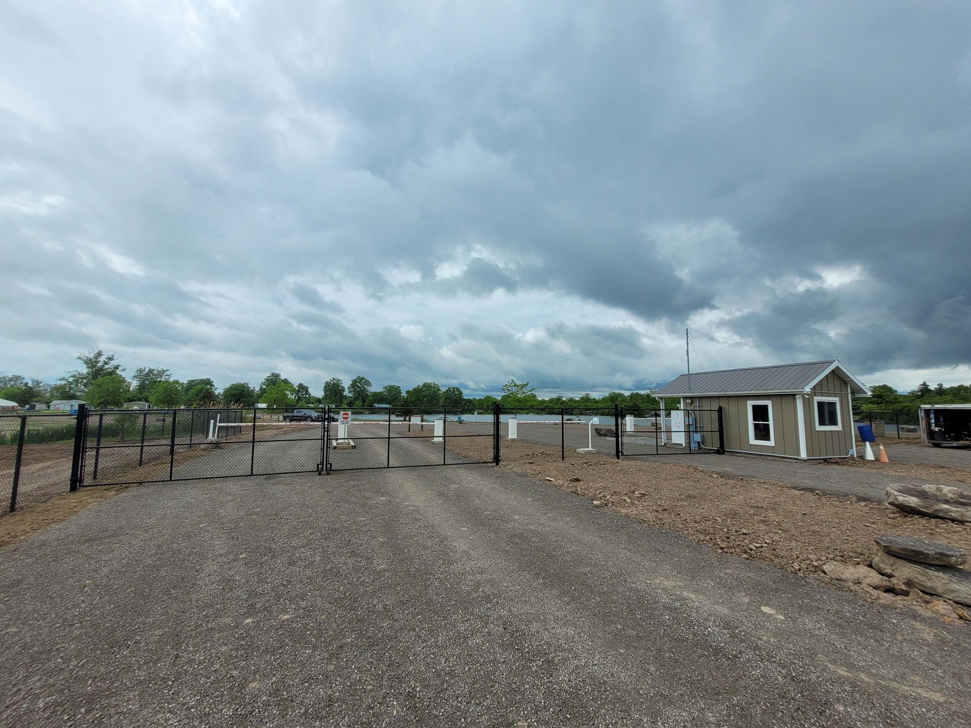 Gravel entrance with a gate, small building, and dark clouds overhead.