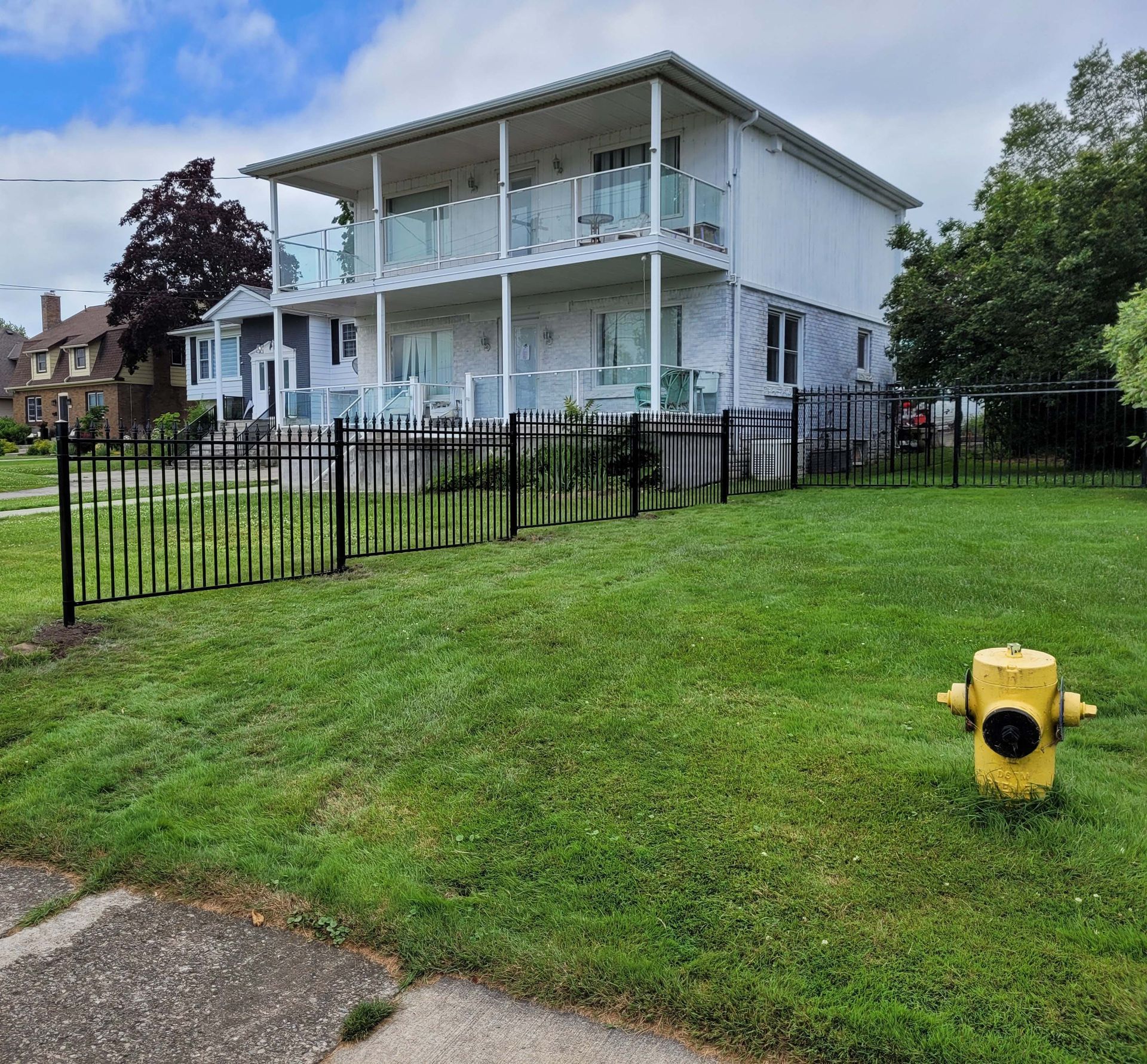 Two-story white house with glass balconies, black fence, and yellow fire hydrant on a grassy lawn.