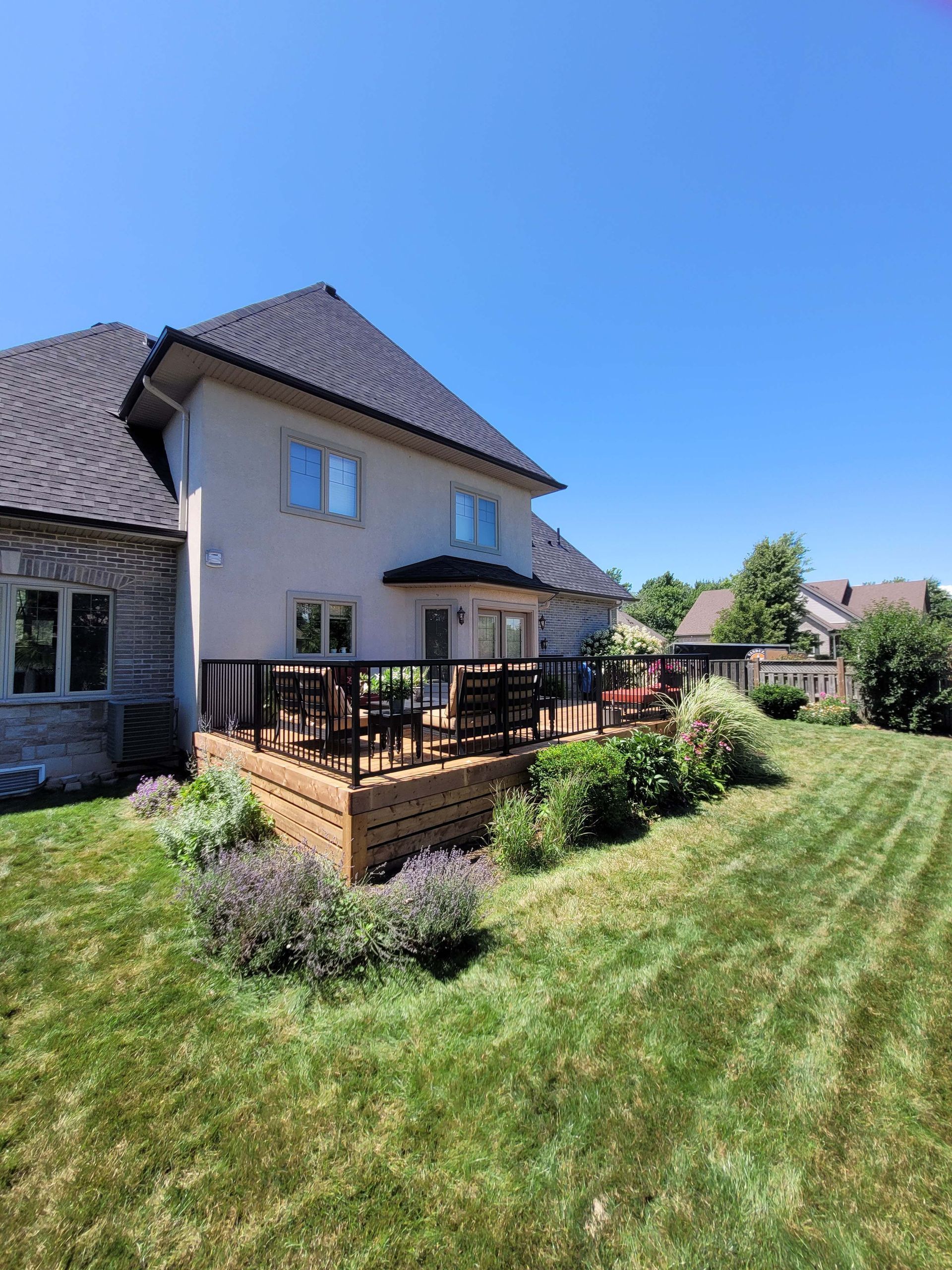 A two-story beige house with a wooden deck, surrounded by a green lawn and blue sky.