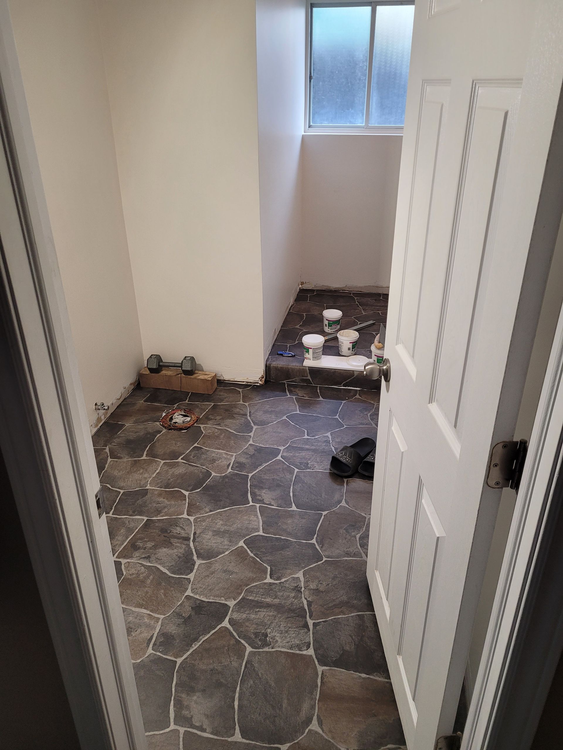 A small room with stone-patterned floor, open white door, small window, and paint supplies.