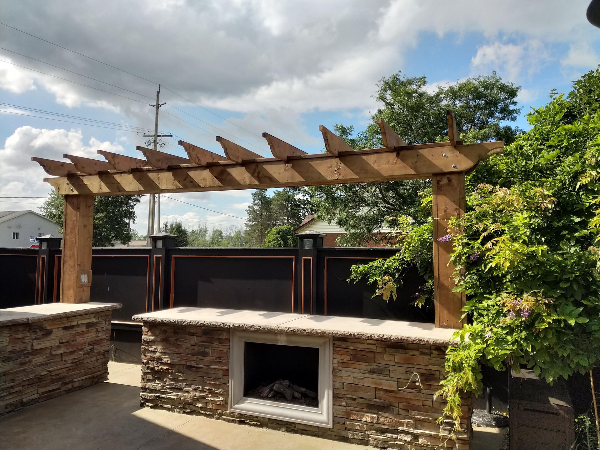 Outdoor kitchen with stone countertop and fireplace, wooden pergola overhead.