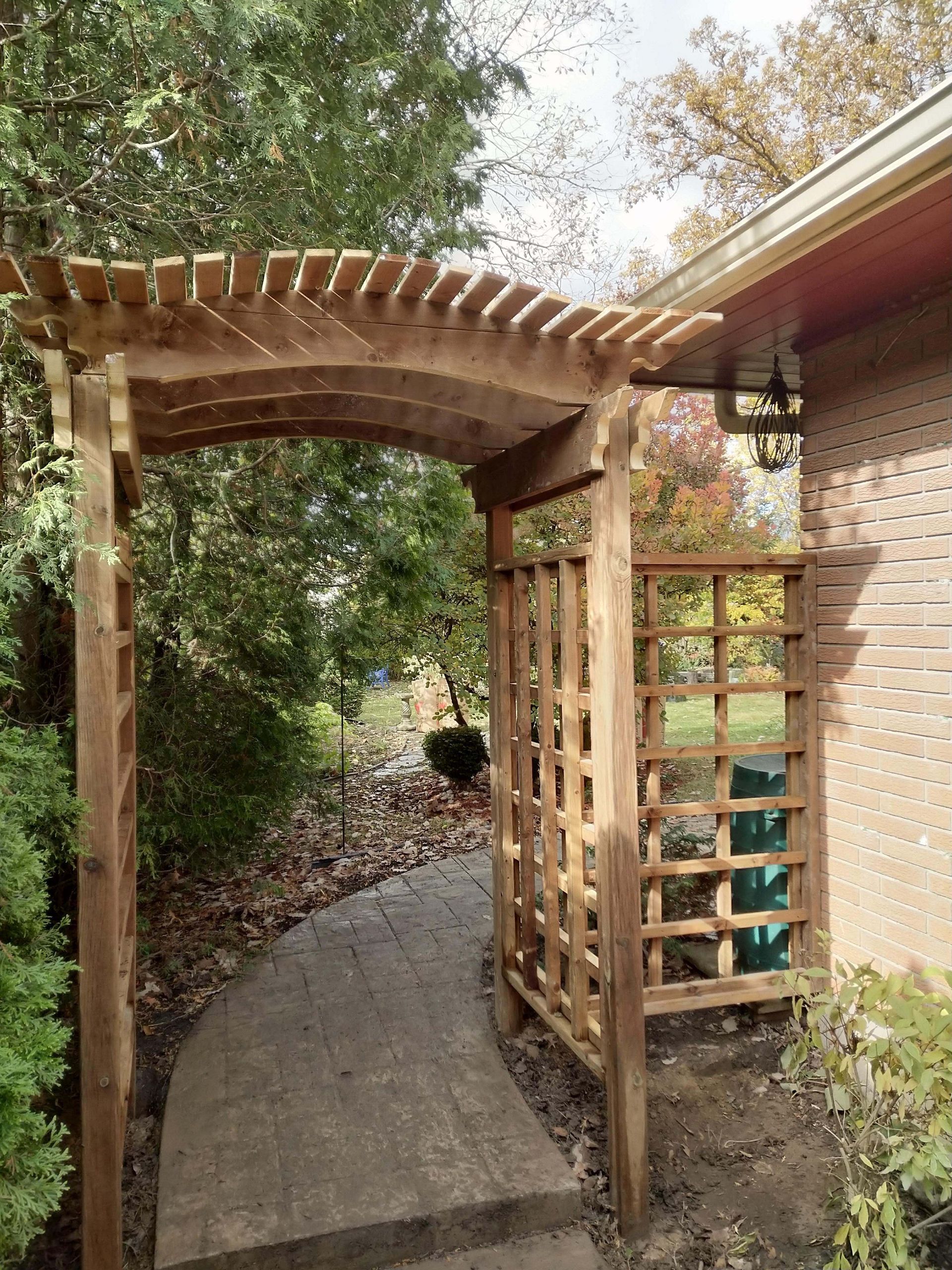 Wooden arbor over a curved walkway, beside a brick building. Lattice wall and greenery in the background.