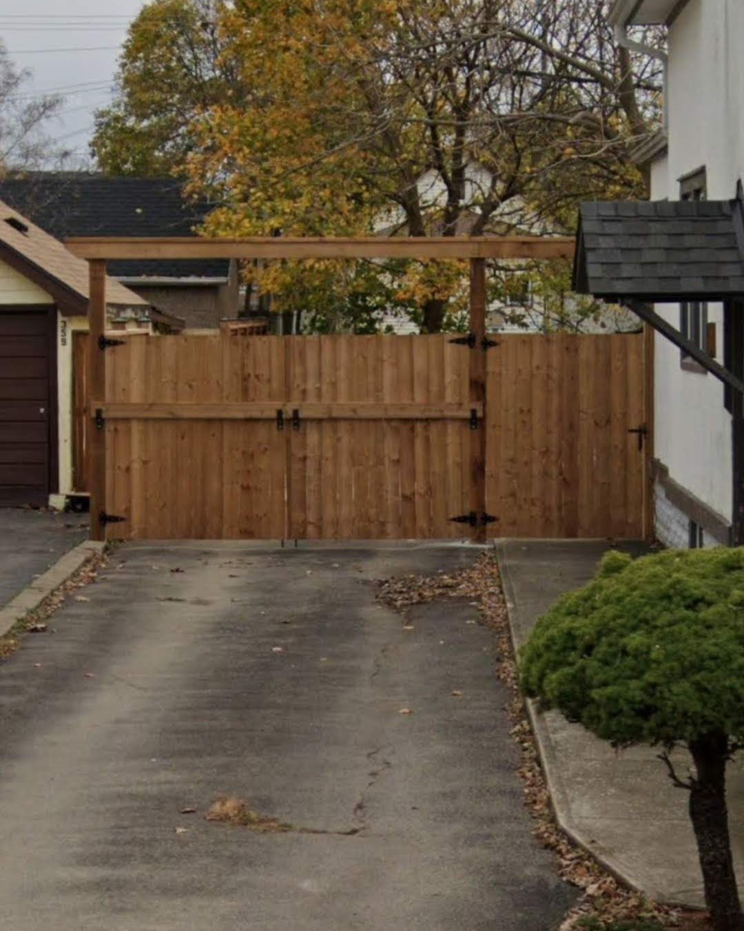 Wooden gate spanning a driveway, secured by black hinges and a latch. A trimmed bush sits to the right.