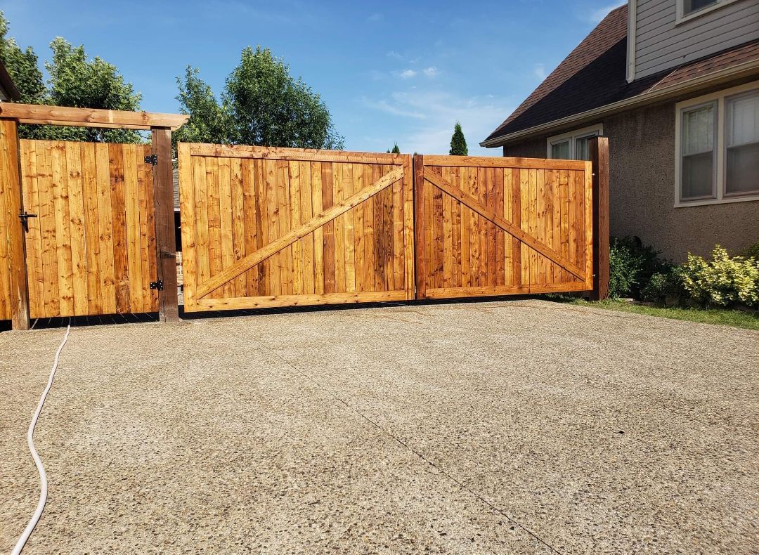 Wooden driveway gate, stained orange, with diagonal supports, gravel ground, near a house.