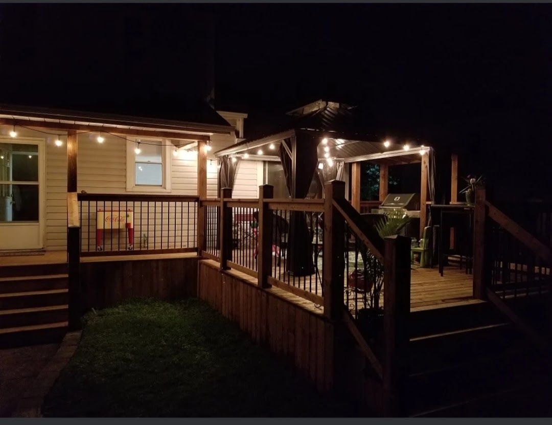 Nighttime view of a wooden deck with string lights and a gazebo, attached to a house.