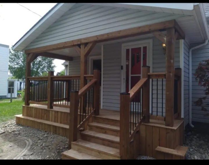 Wooden porch with steps, railing, and overhead structure, attached to a light-colored house with a red door.