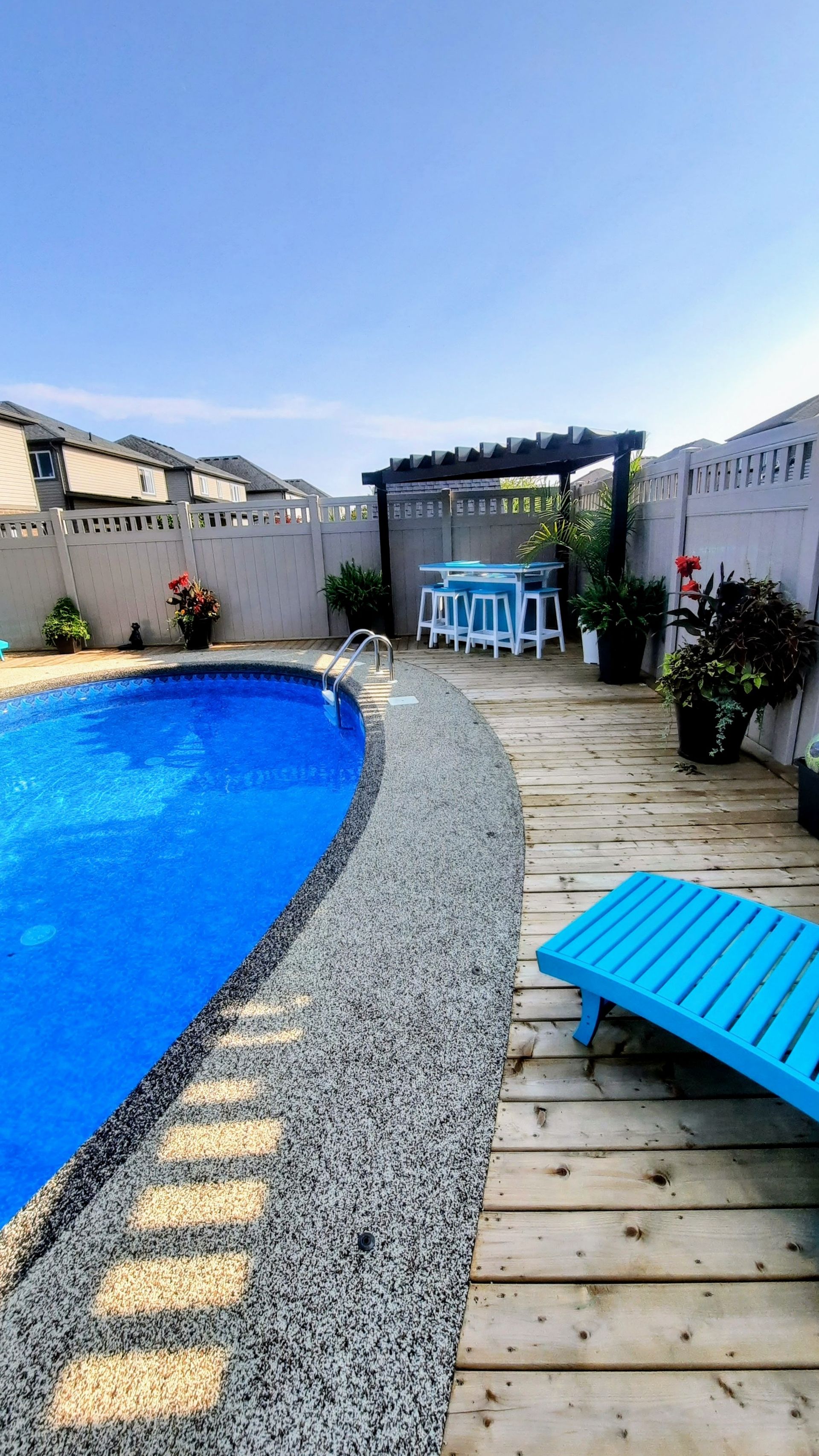 Blue pool and deck with a table set under a pergola on a sunny day.